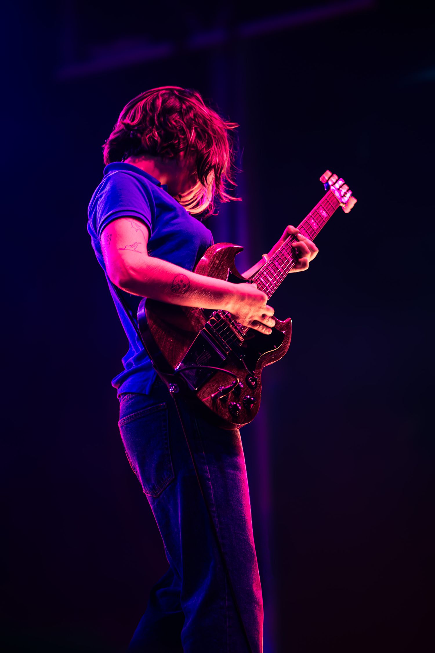 Guitarist performing on stage under purple and pink stage lights, playing an electric guitar with intense focus.