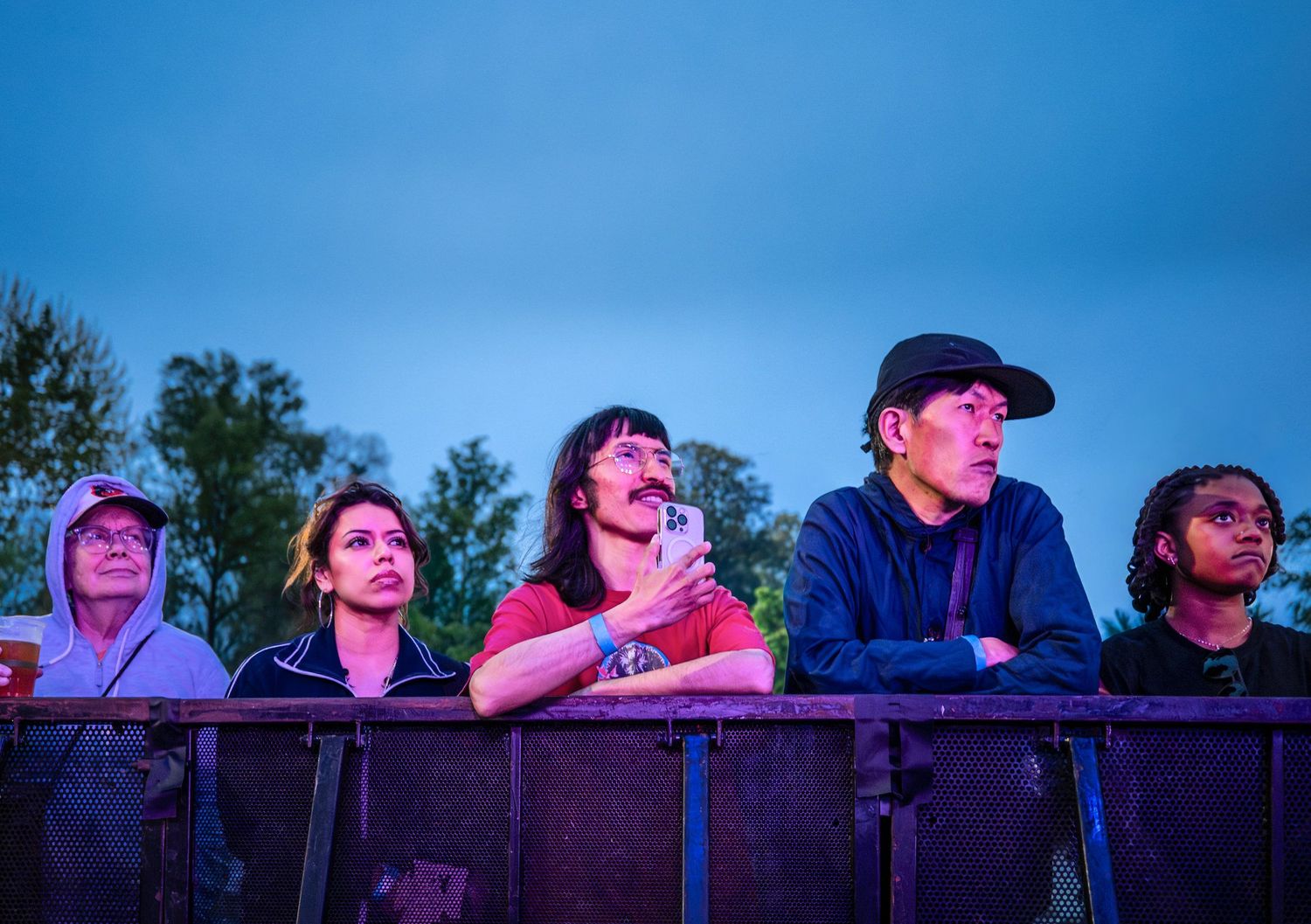 Group of young festival attendees looking toward a stage at dusk under a blue sky with trees behind.