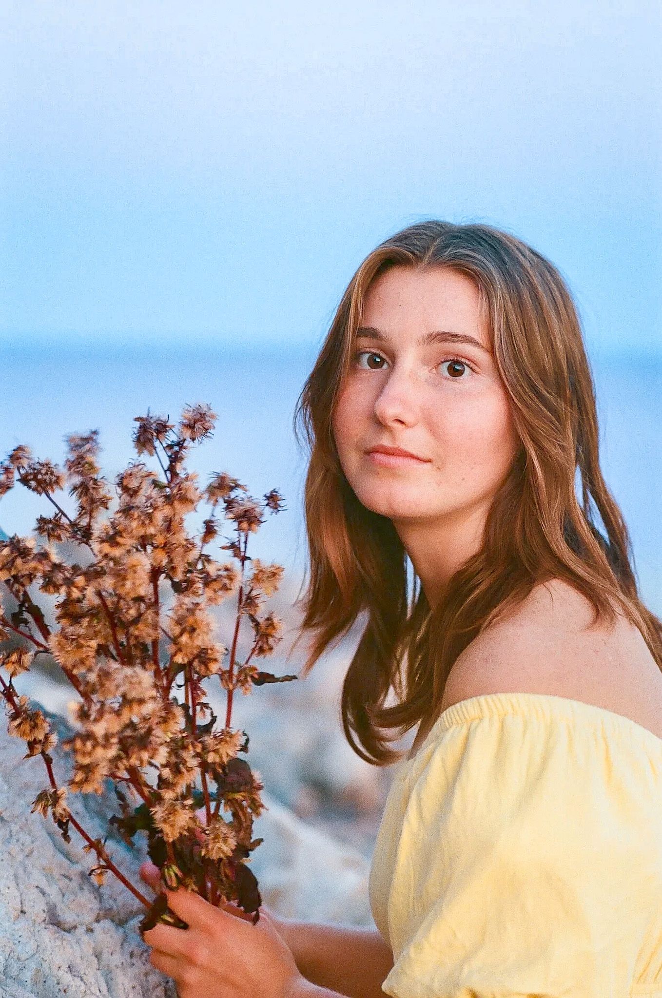 Young woman with auburn hair in yellow off-shoulder dress holds dried wildflowers near a serene blue ocean at dusk.