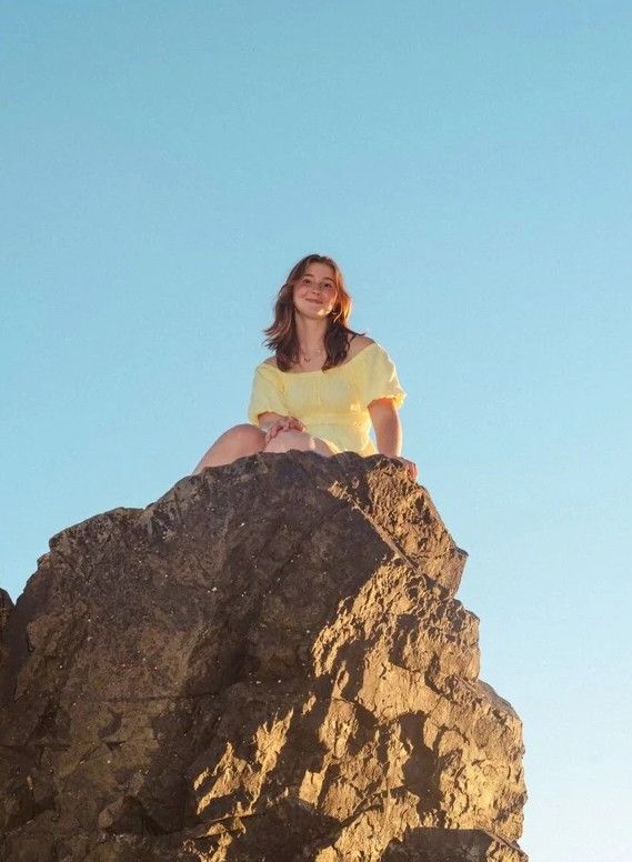 Young woman in yellow dress sitting atop a rocky boulder against a clear blue sky, smiling in golden sunlight.