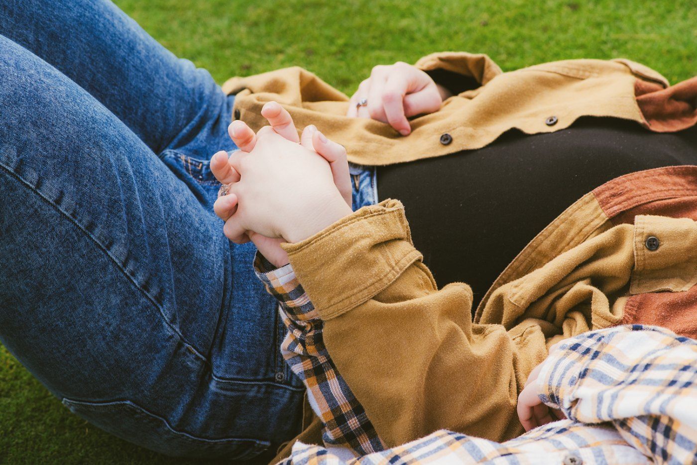 Pregnant woman lying on grass with hands clasped over baby bump, wearing jeans and brown jacket outdoors.