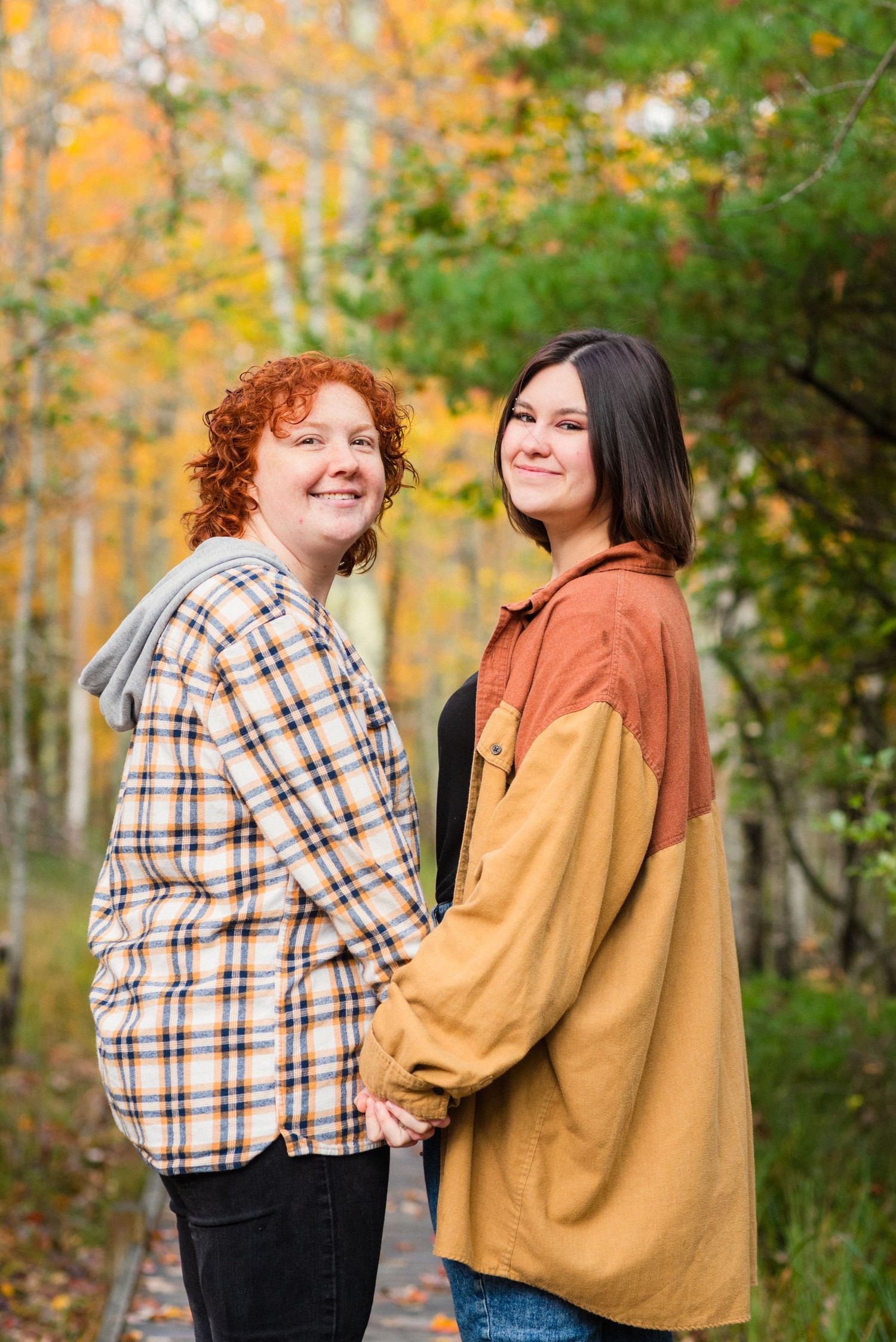 Two women holding hands on a tree-lined path surrounded by vibrant autumn foliage in fall colors.