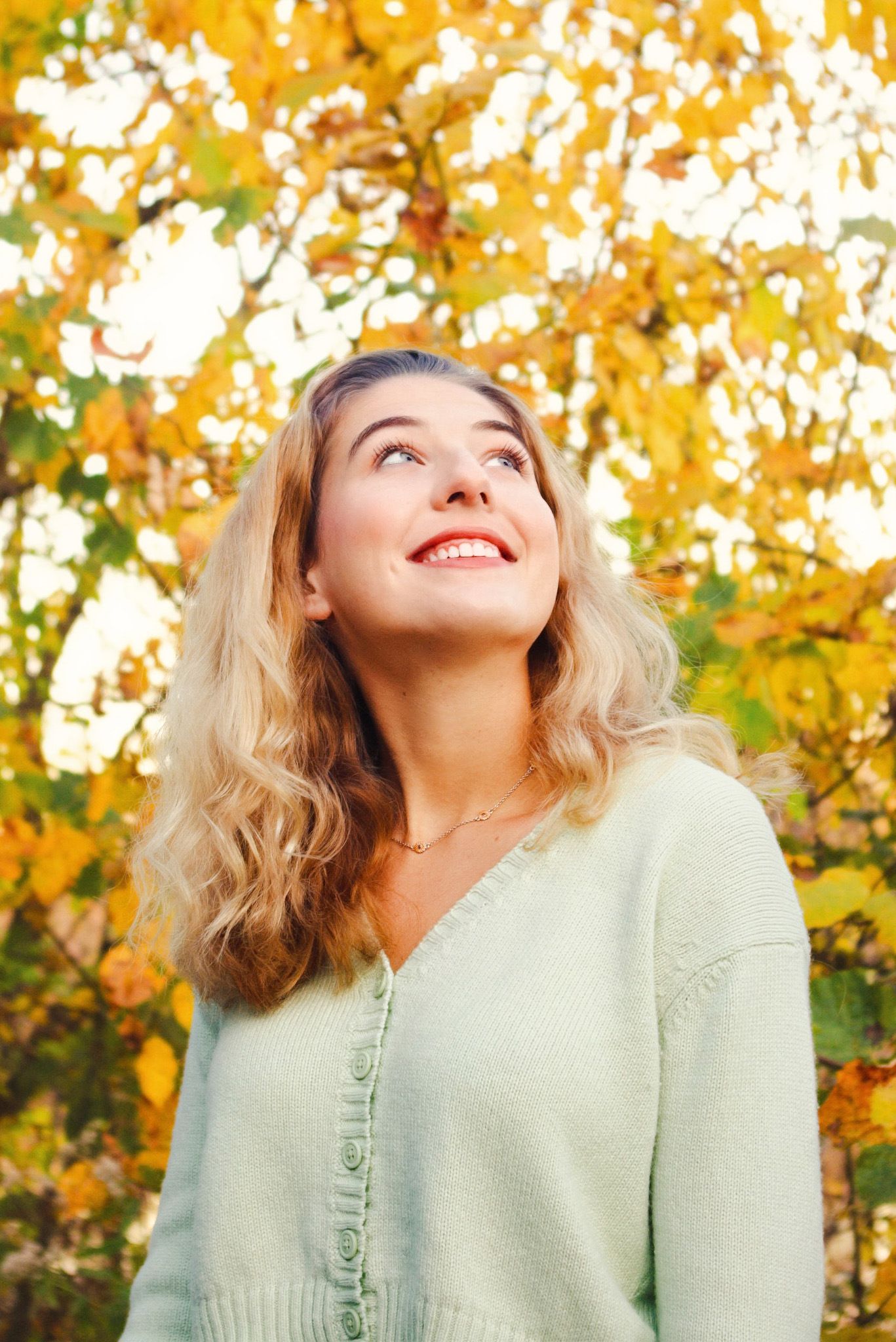 Smiling blonde woman in mint green sweater looking up at colorful autumn yellow leaves in outdoor fall setting.