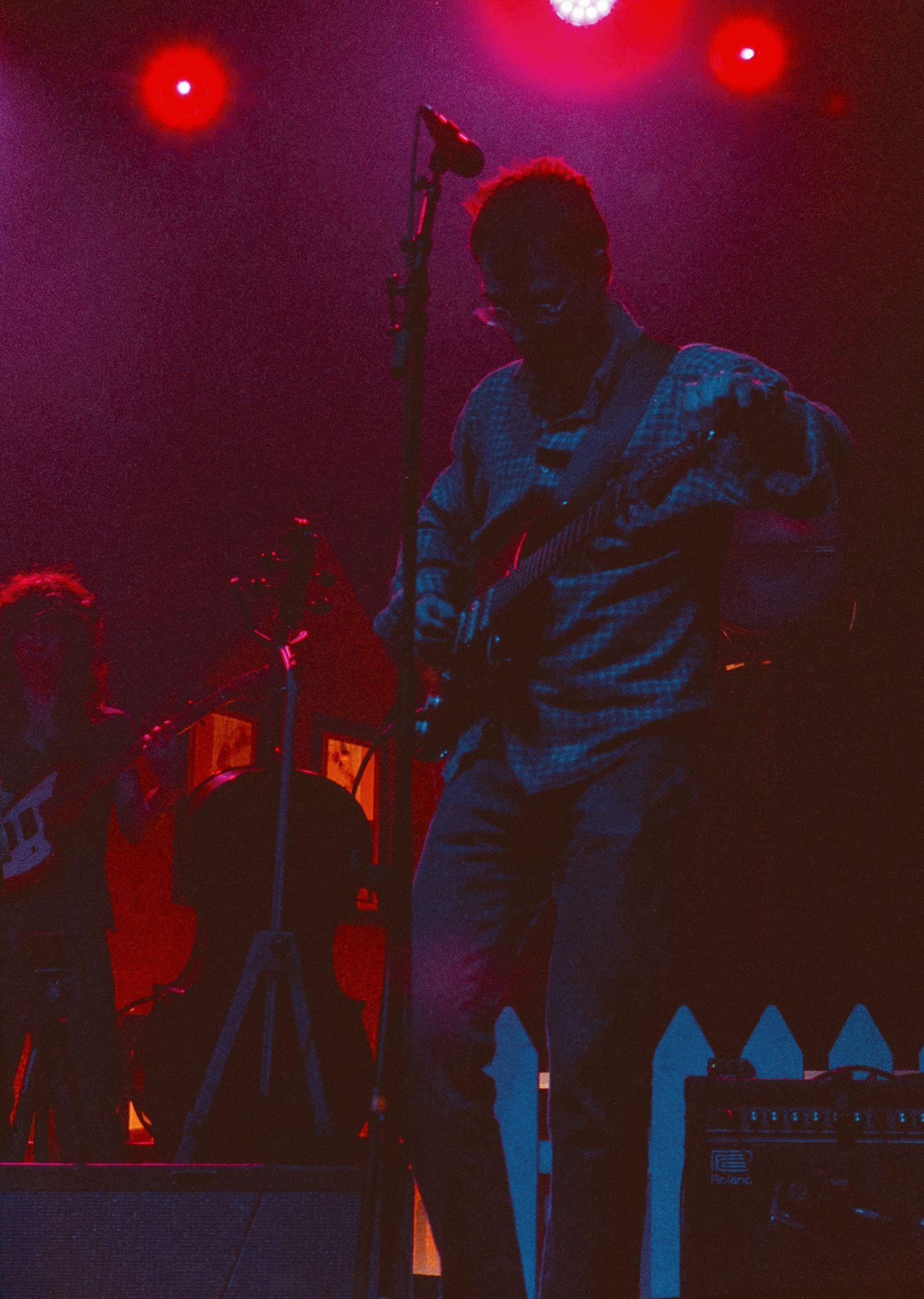Silhouetted guitarist performing under dramatic red stage lighting with a white picket fence prop on stage.