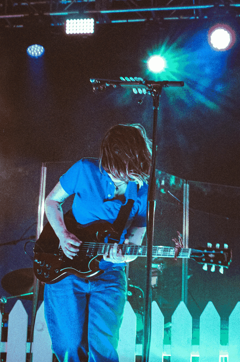 Guitarist performing on stage under blue stage lights with a black electric guitar near a microphone stand.