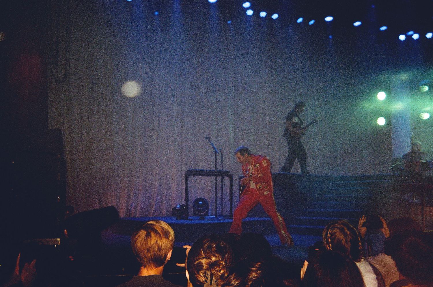 Two performers on a dimly lit stage, one in a red outfit dancing near keyboard equipment.