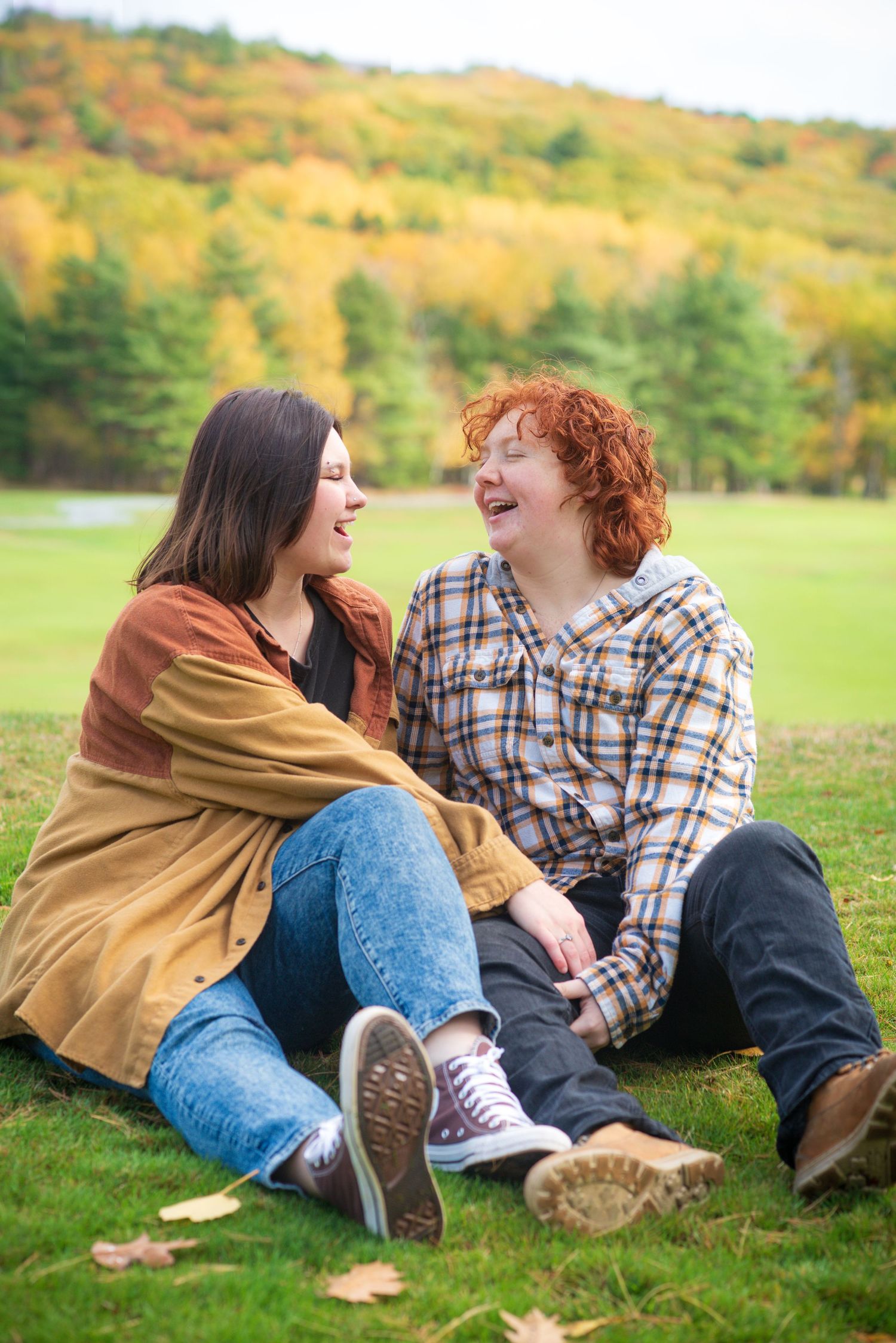 Two friends laughing together on grass with colorful autumn foliage hillside backdrop on a fall day.
