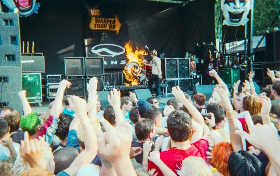 Band performing on Warped Tour stage with pyrotechnics as excited crowd raises hands in the air.