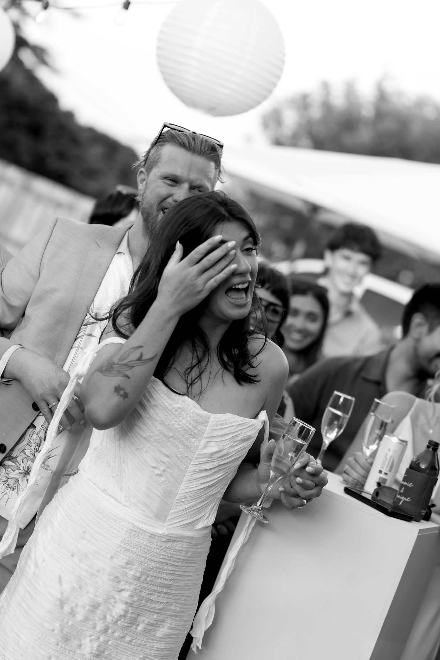 A bride in a white dress laughs joyfully at an outdoor wedding reception while holding a champagne glass.