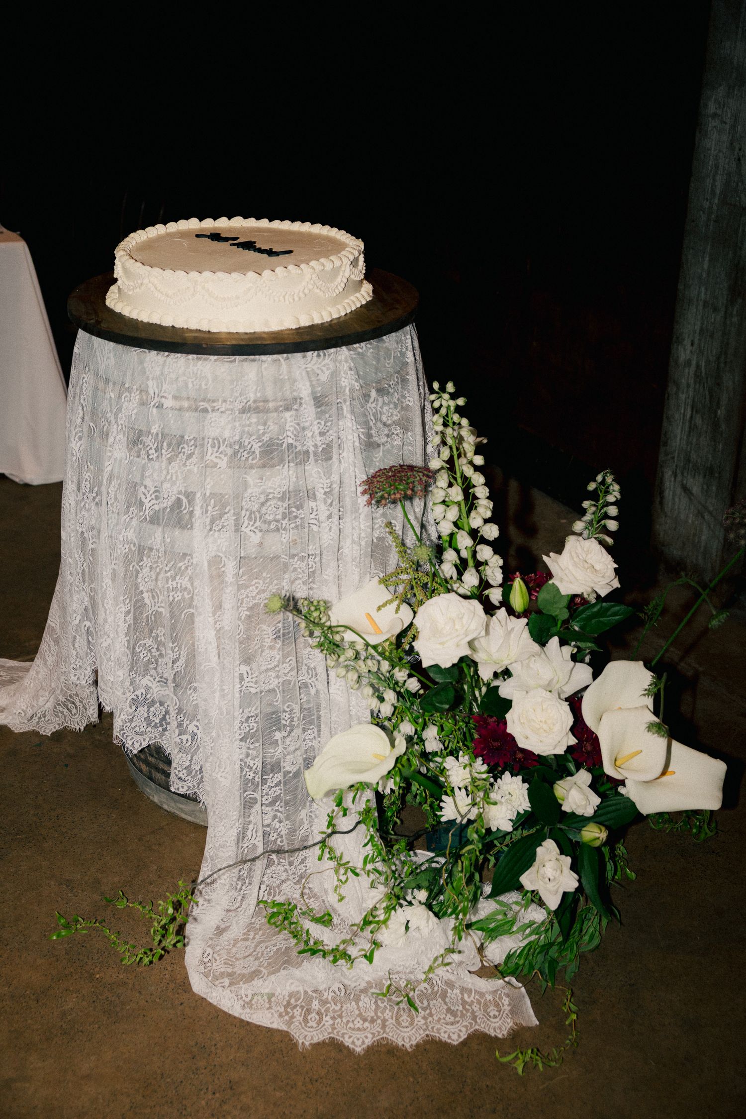 Wedding cake displayed on a lace-draped barrel surrounded by white calla lilies and floral arrangements.