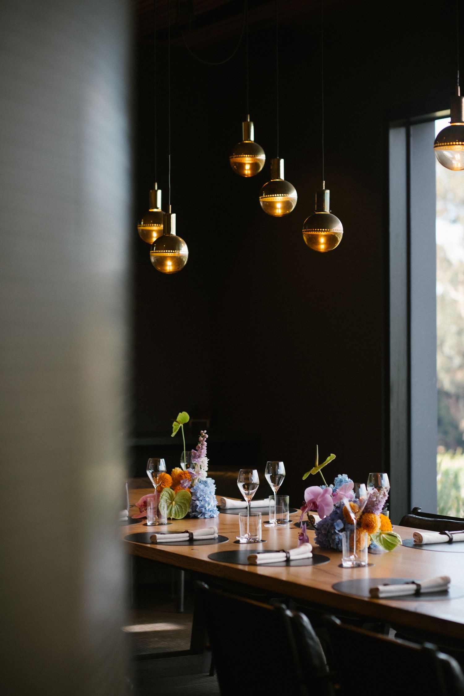 Dark moody wedding reception table with gold pendant lights, floral centerpieces, and glassware settings.