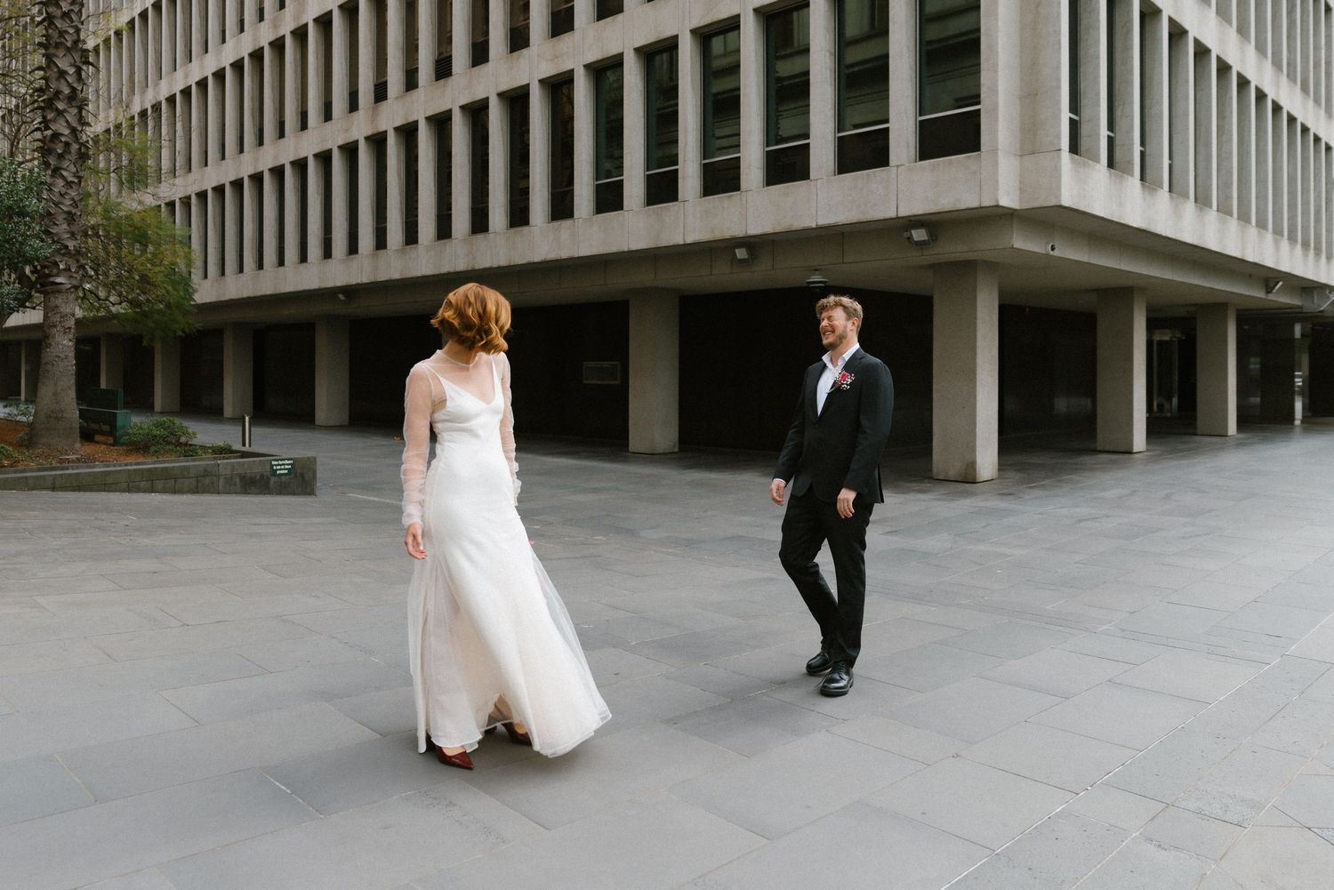 Bride in a white gown and groom in a tuxedo walk toward each other in an urban plaza.
