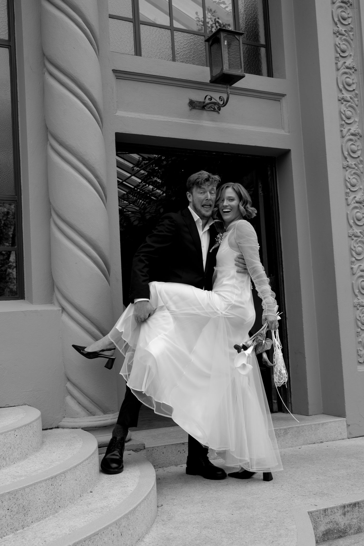 Black and white photo of groom lifting bride on steps outside a grand building entrance.