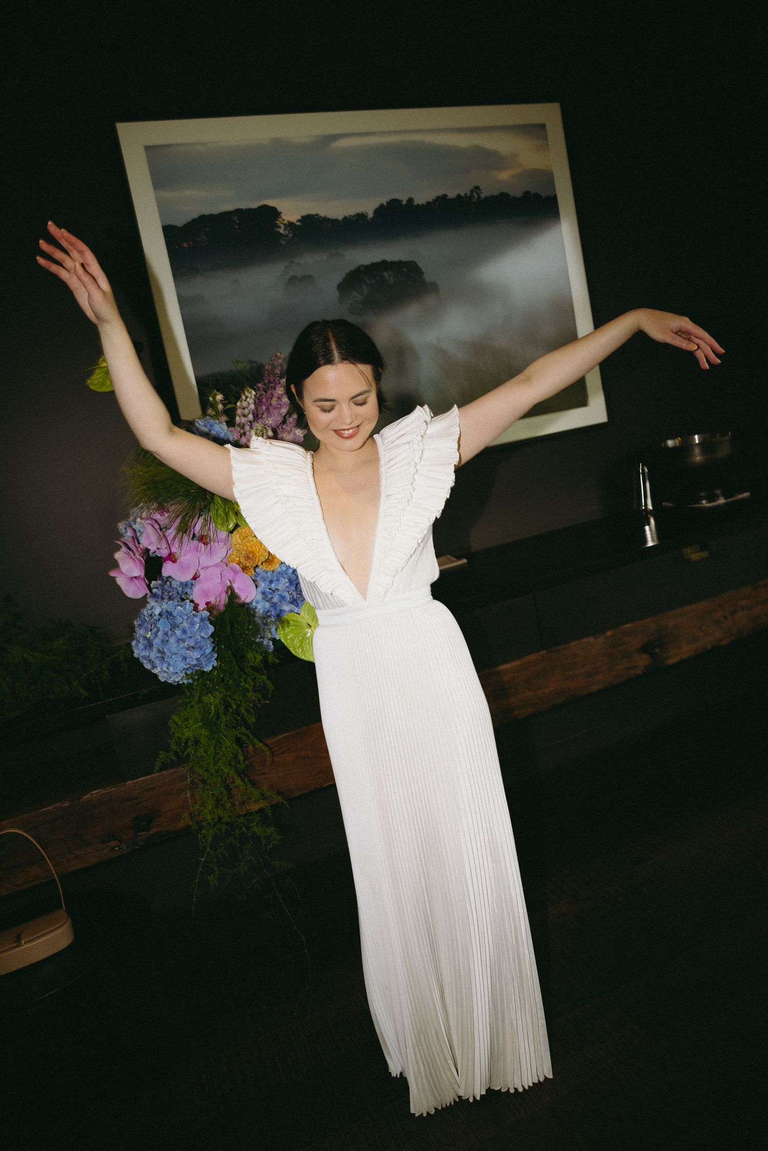 Bride in white pleated deep V-neck gown dancing with arms raised beside colorful floral arrangement.