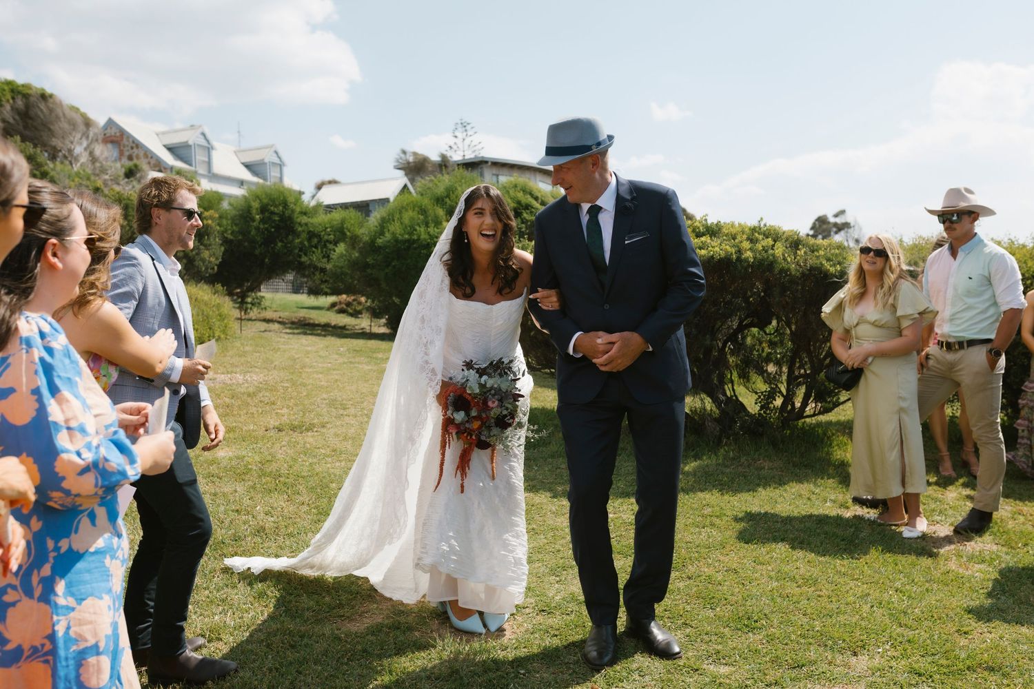 Bride and groom walk down outdoor aisle at rustic garden wedding ceremony, guests watching, bride holding bouquet.