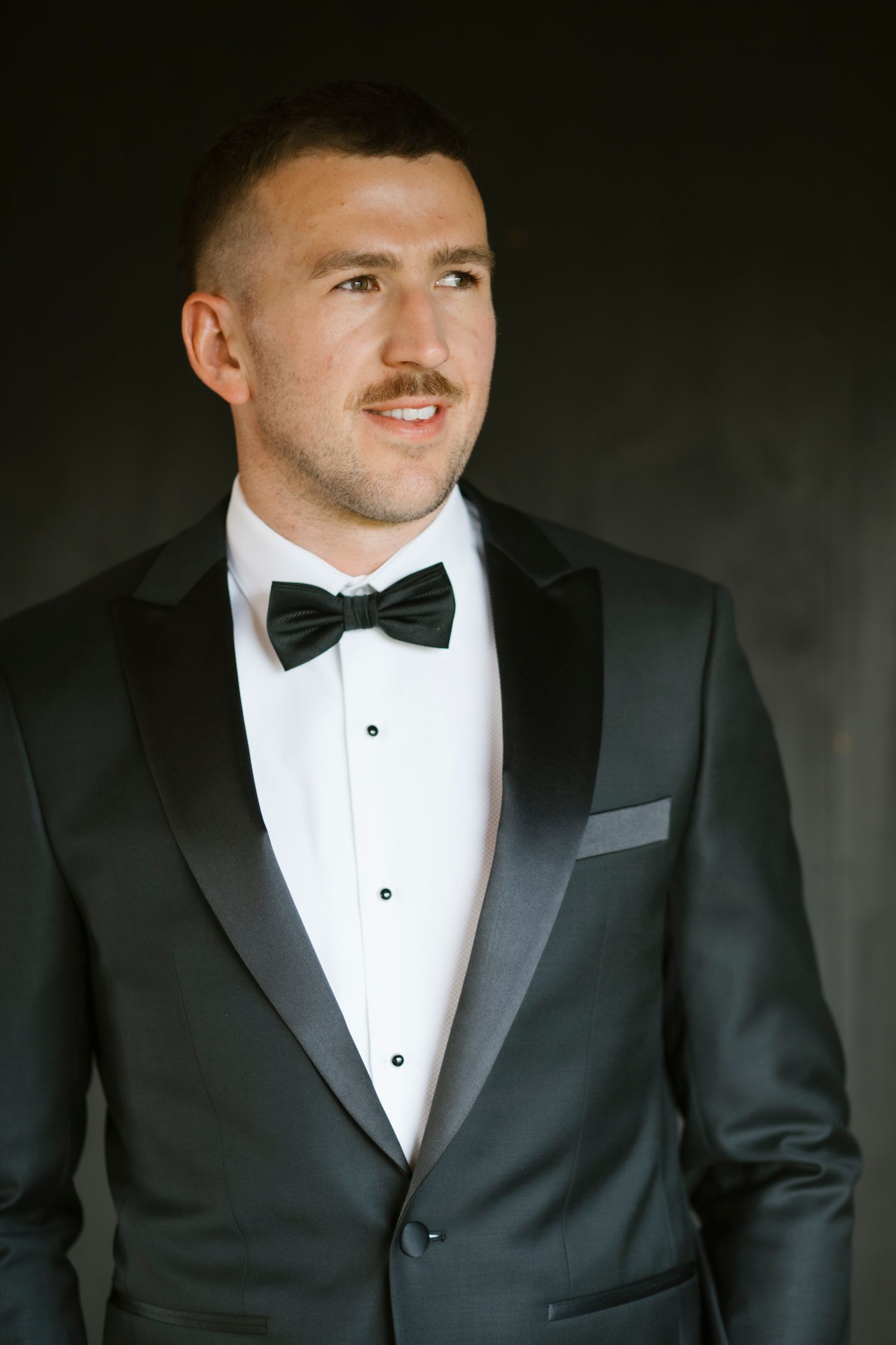 Handsome groom in a classic black tuxedo with bow tie posing against a dark background.