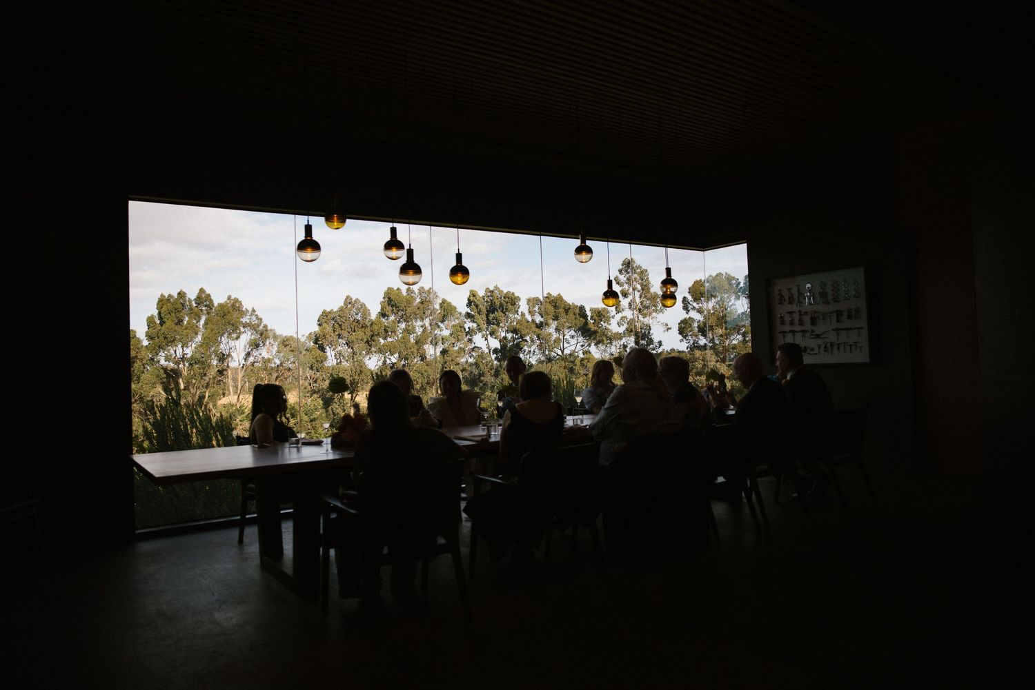 Wedding guests dining in silhouette against large window with scenic bush landscape view.