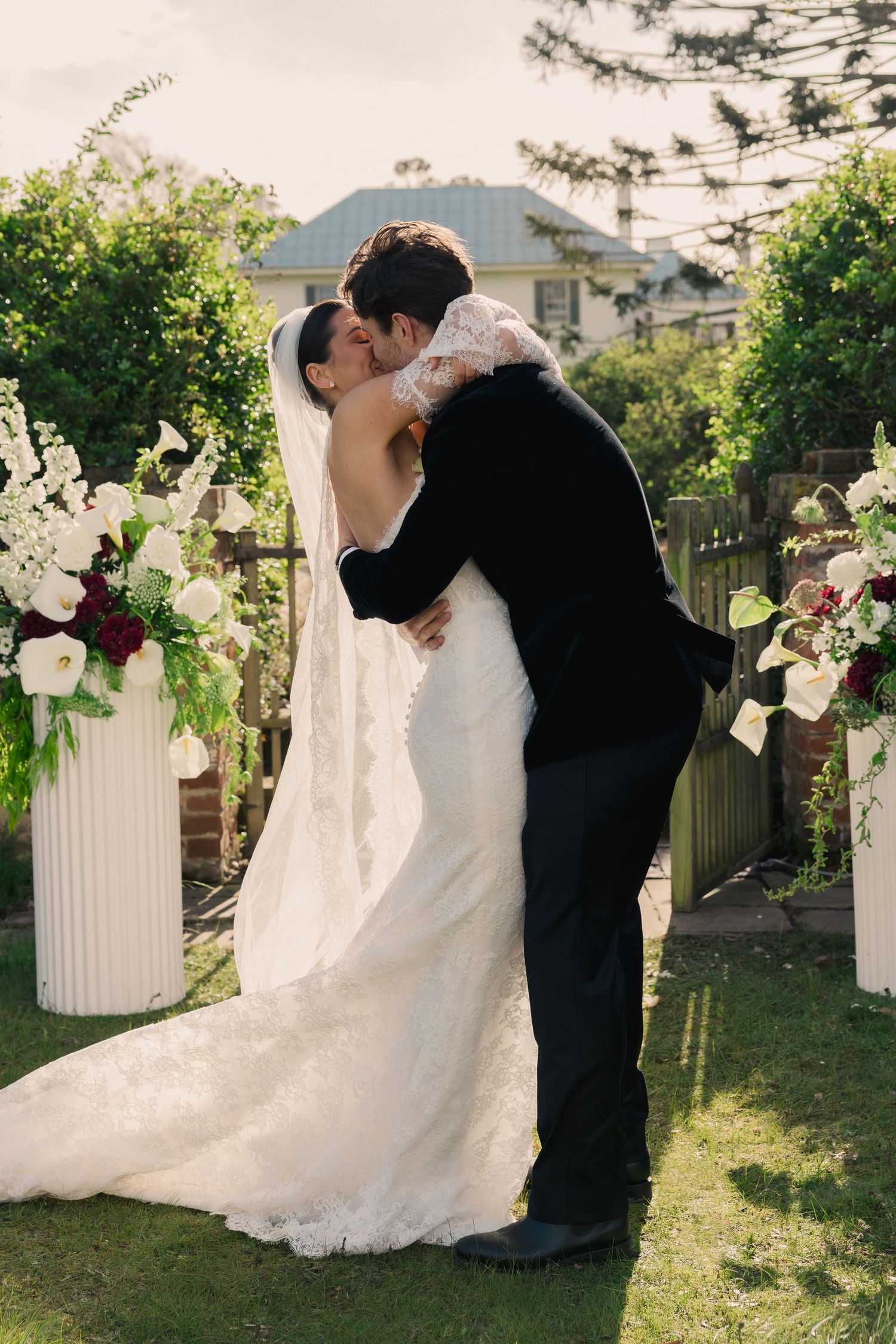 Bride and groom share their first kiss at an outdoor garden wedding ceremony surrounded by white floral arrangements.