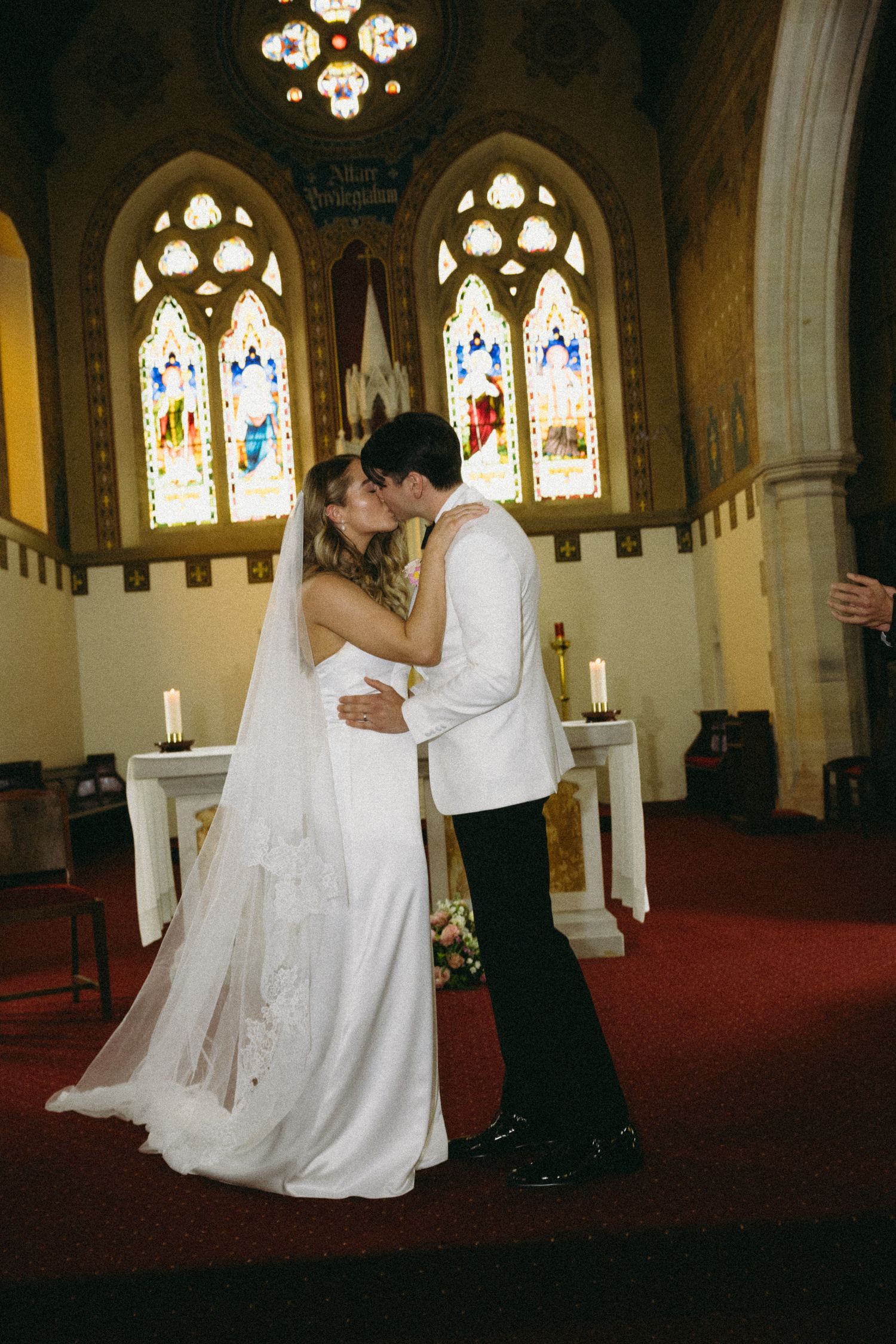 Bride and groom sharing first kiss at altar in ornate church with stained glass windows.