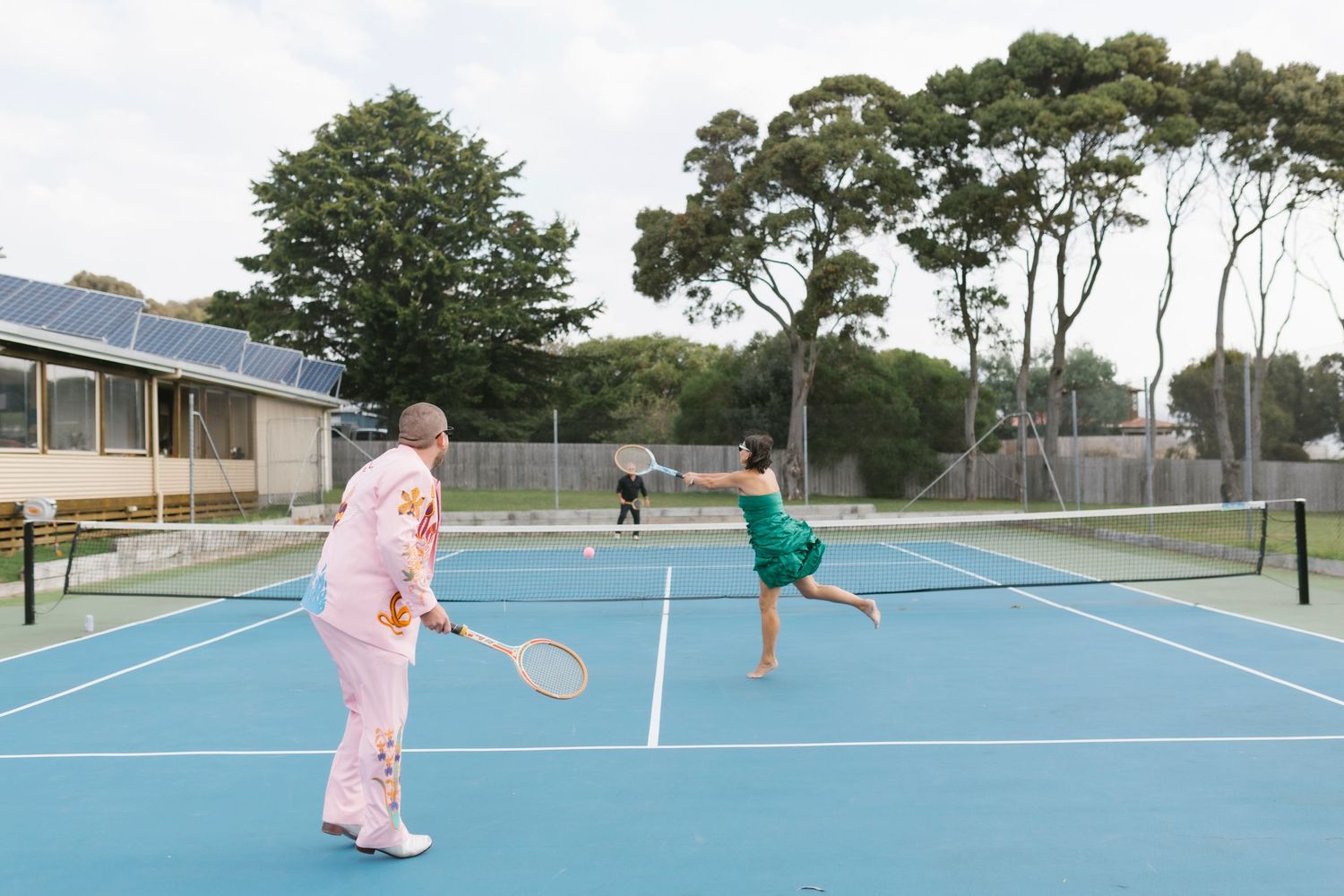 Wedding guests playing tennis in colorful outfits on blue court surrounded by trees.