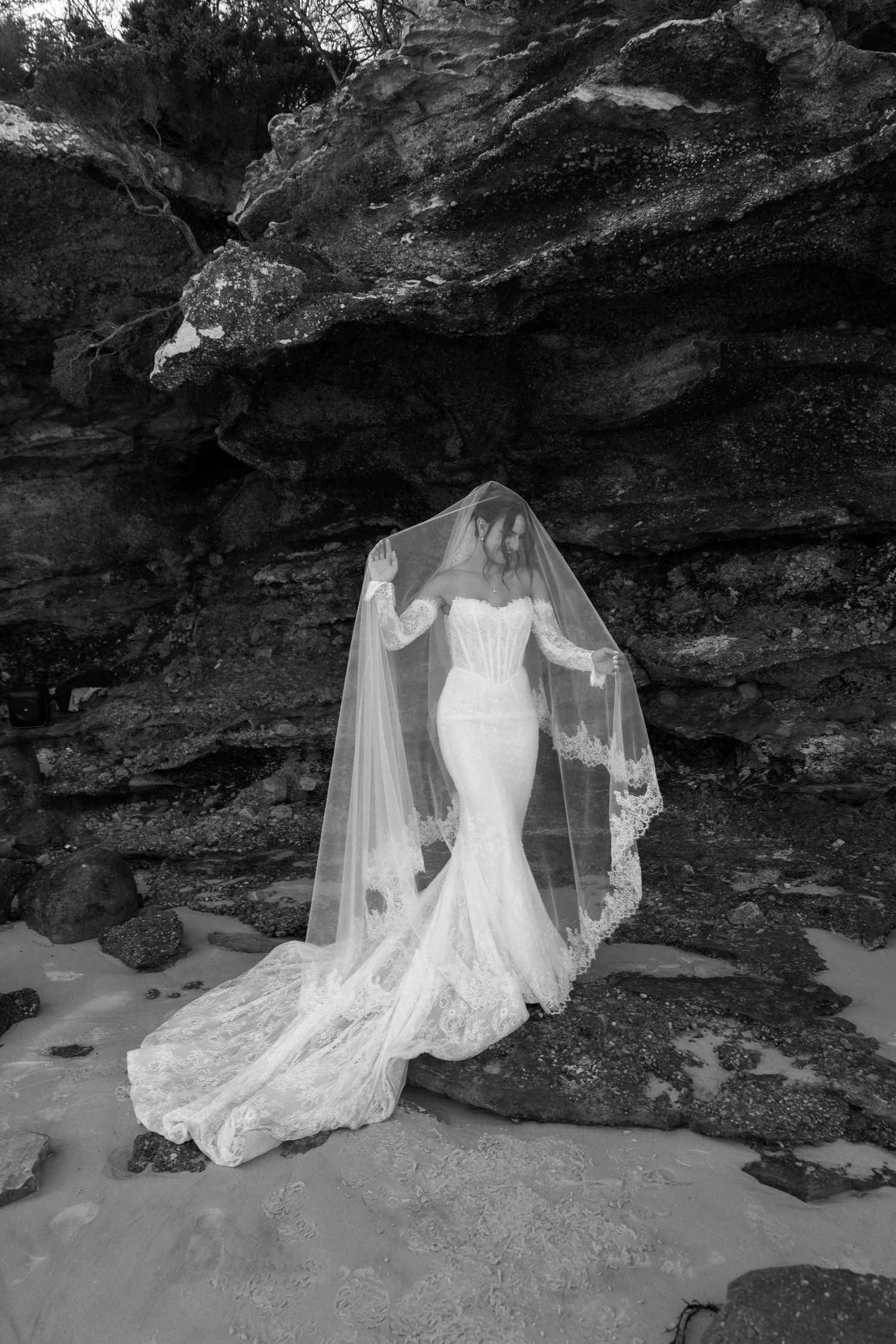 Black and white photo of bride in mermaid lace gown lifting veil against dramatic rock face.