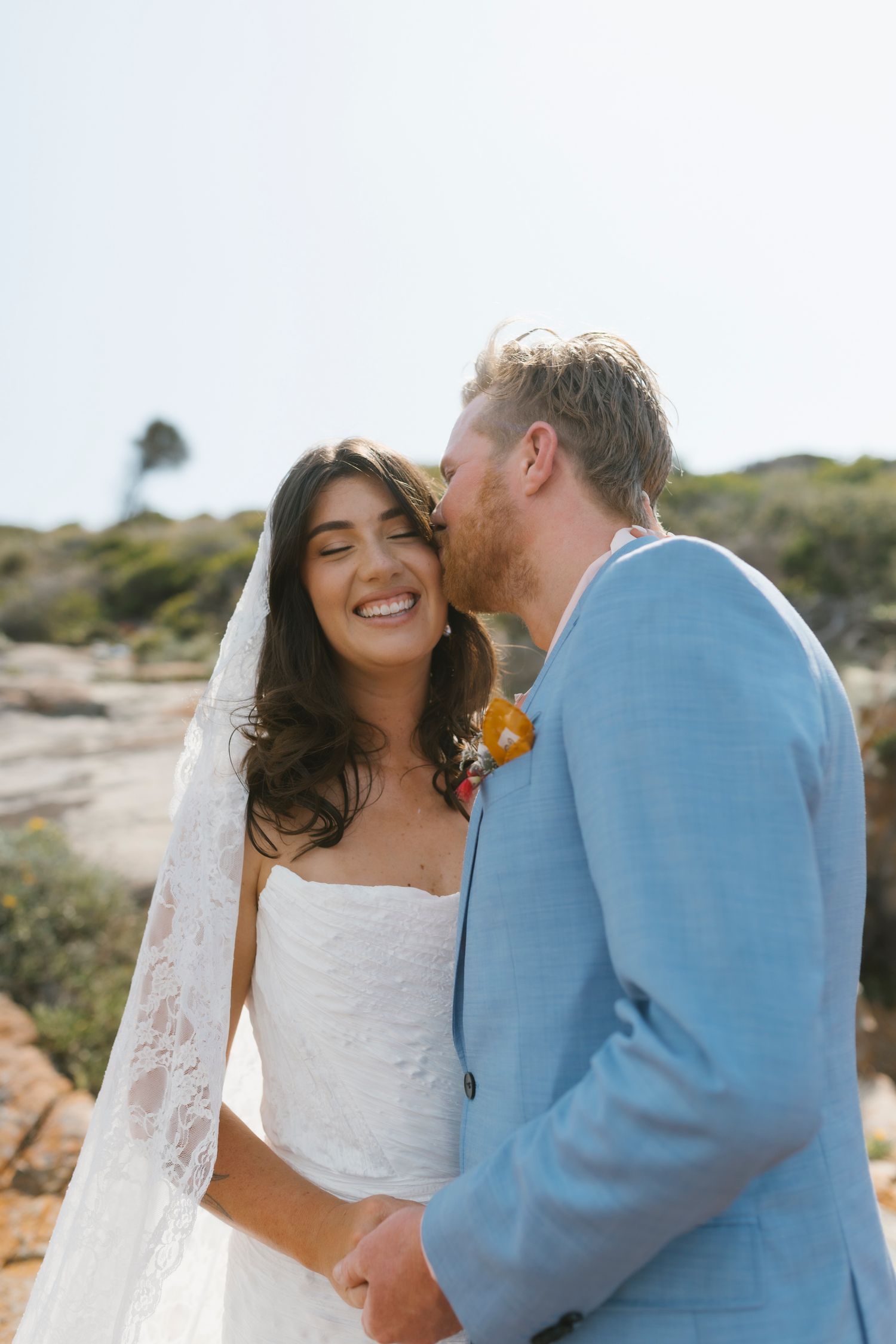 Bride in white gown and veil smiles as groom in light blue suit kisses her cheek outdoors.