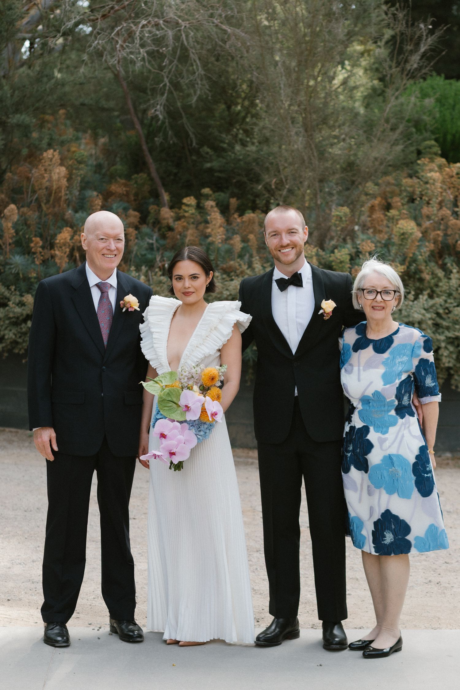 Wedding couple posing with parents outdoors, bride in ruffled white gown holding colorful bouquet.