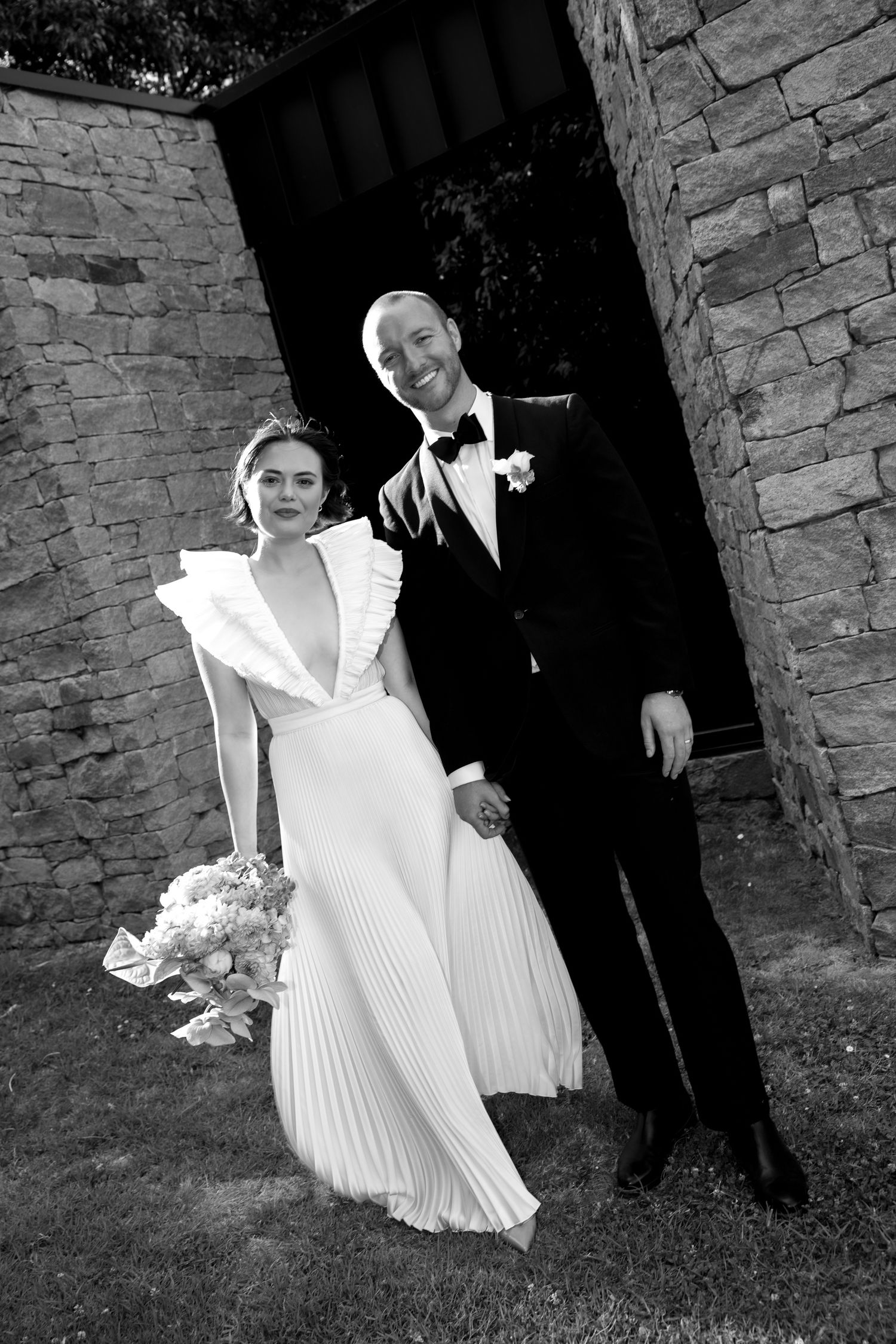 Black and white portrait of elegant bride and groom standing before stone archway at wedding venue.