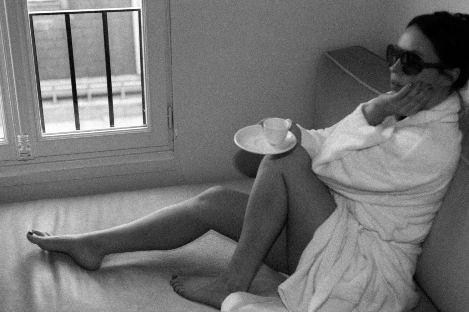 Black and white photo of a bride in a white robe and sunglasses relaxing by a window with a cup of tea.