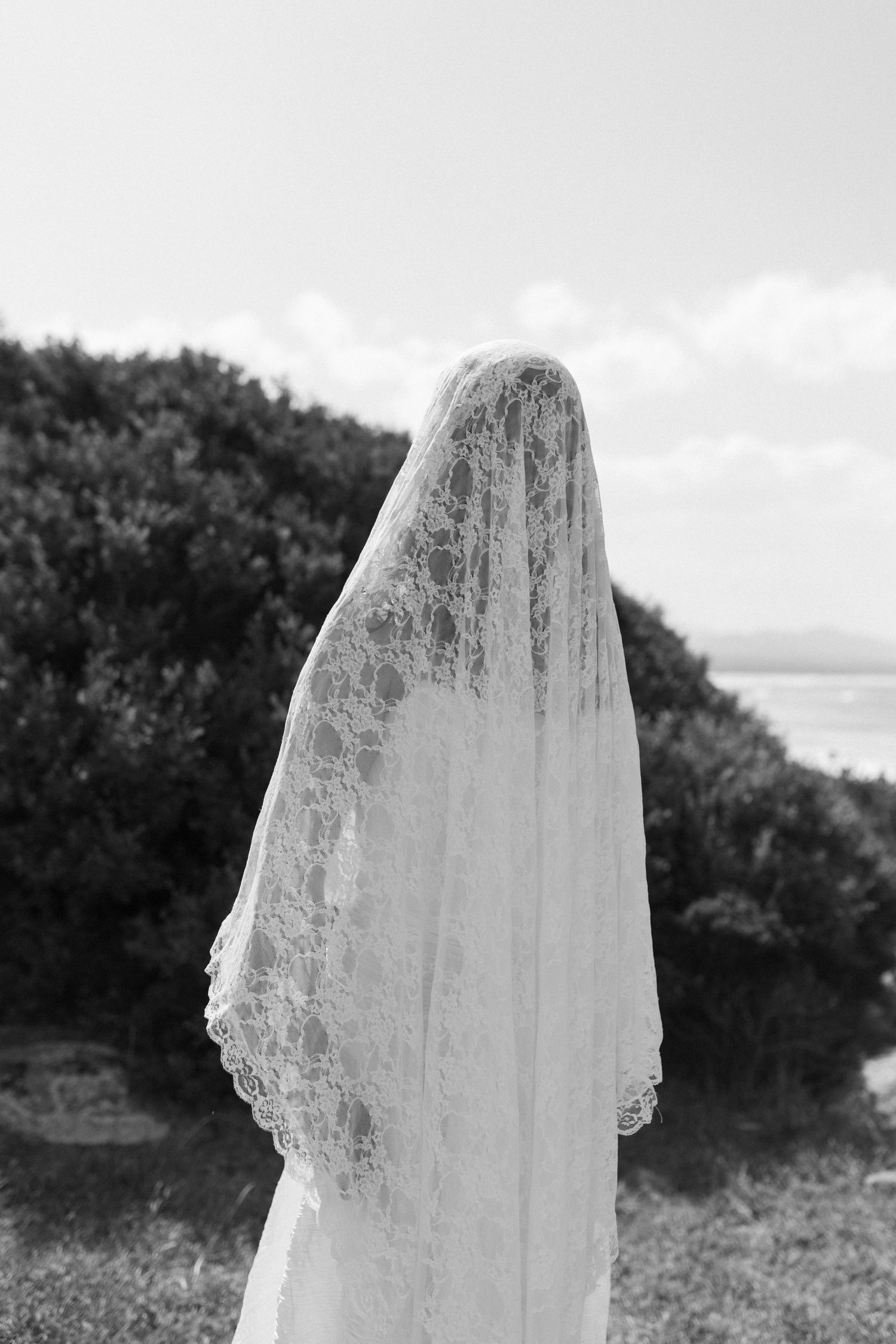 Black and white rear view of a bride in a white gown with an elegant lace veil outdoors.
