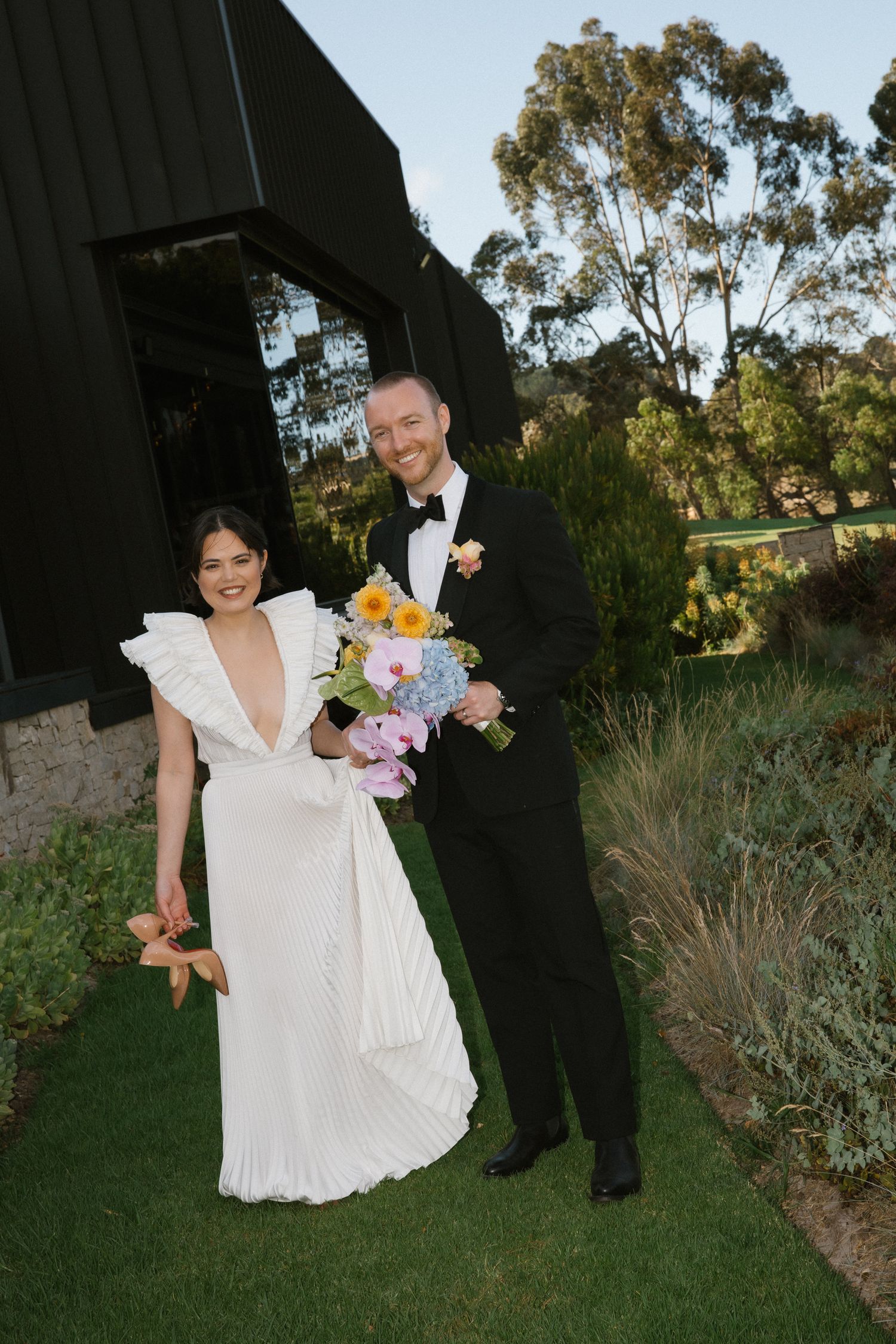Smiling bride and groom posing outdoors with colorful bouquet beside black barn building.