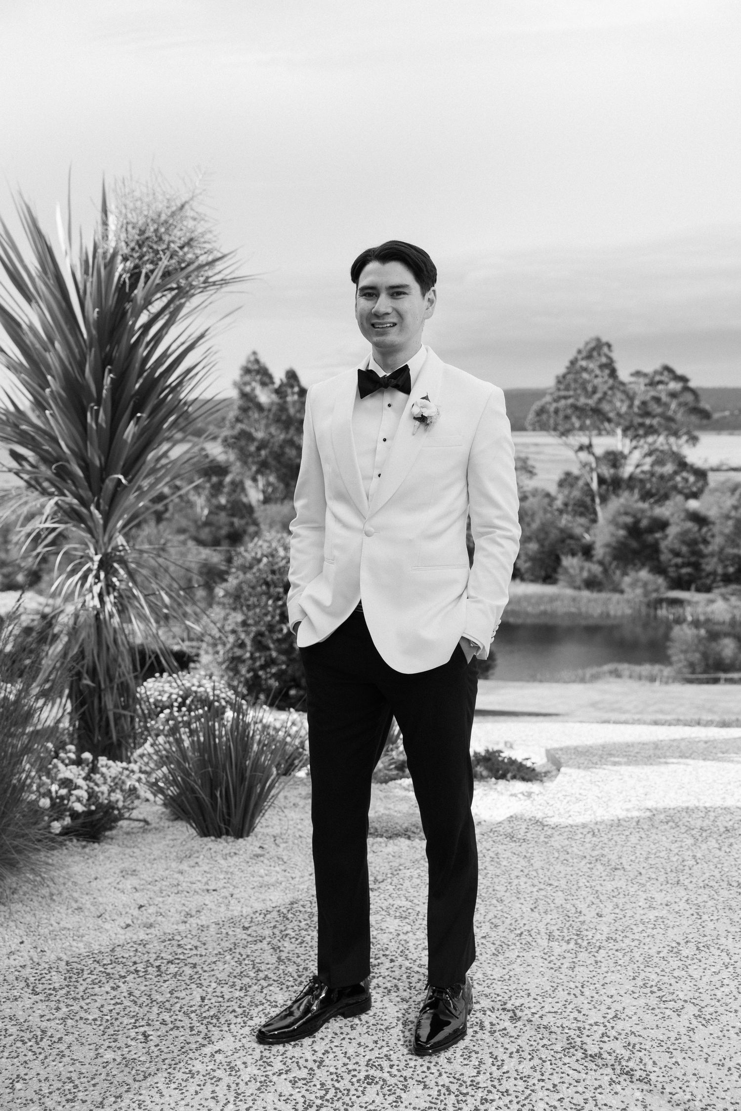 Dapper groom in white tuxedo jacket and black bow tie posing outdoors in black and white photo.
