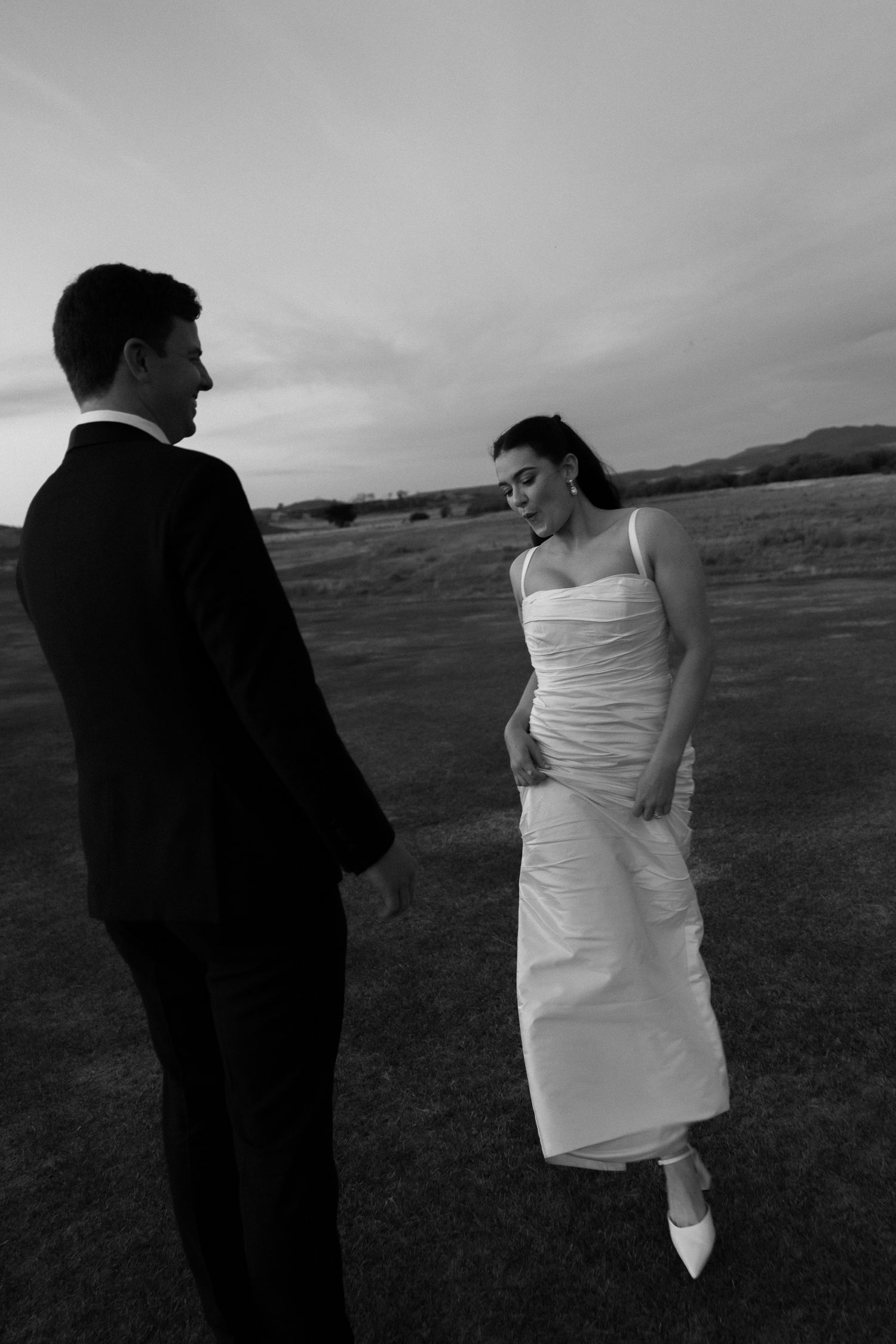 Black and white photo of bride and groom sharing a laugh in an open field at dusk.