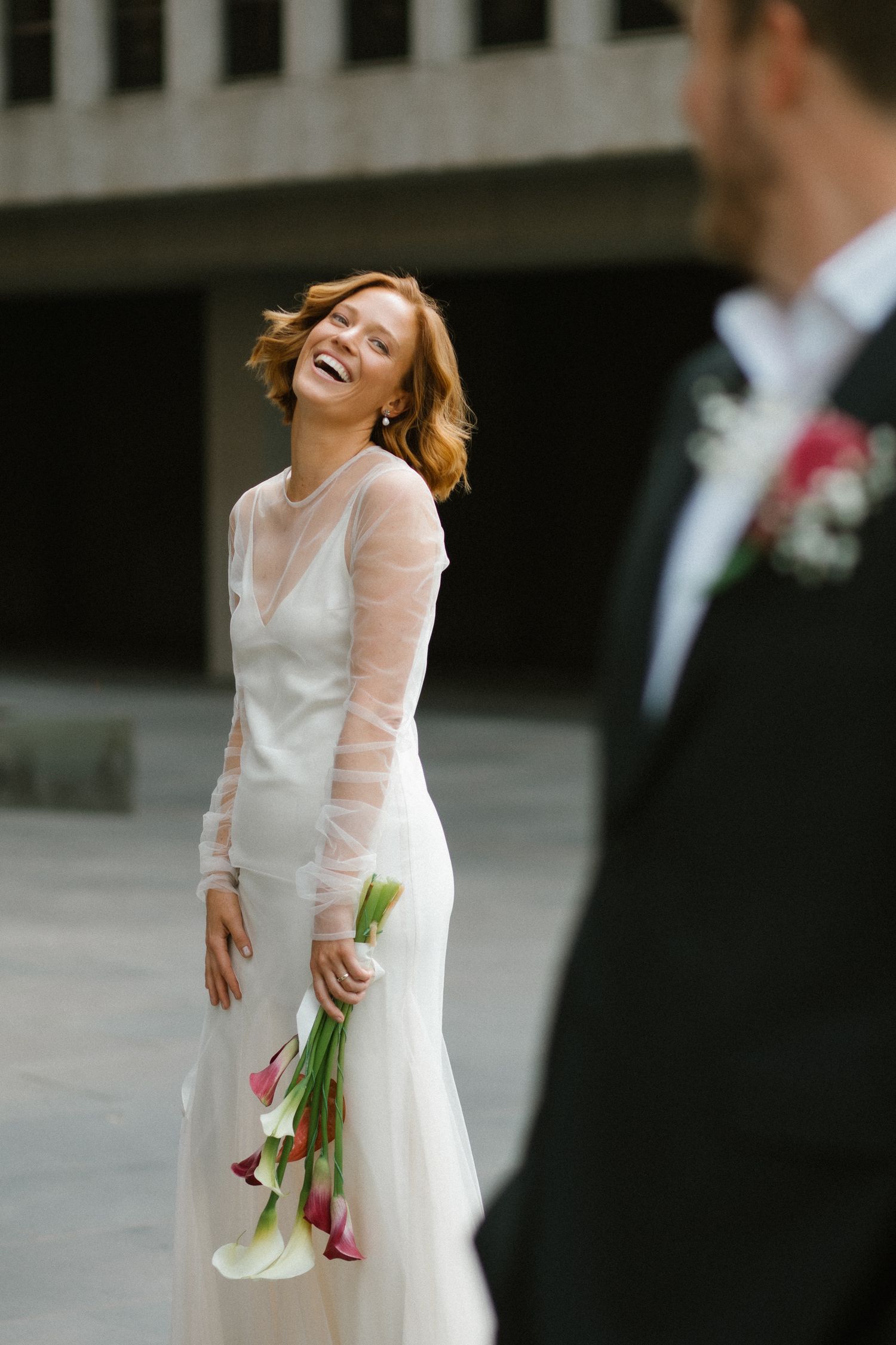 Laughing bride in white sheer-sleeved gown holding calla lily bouquet with groom in foreground.