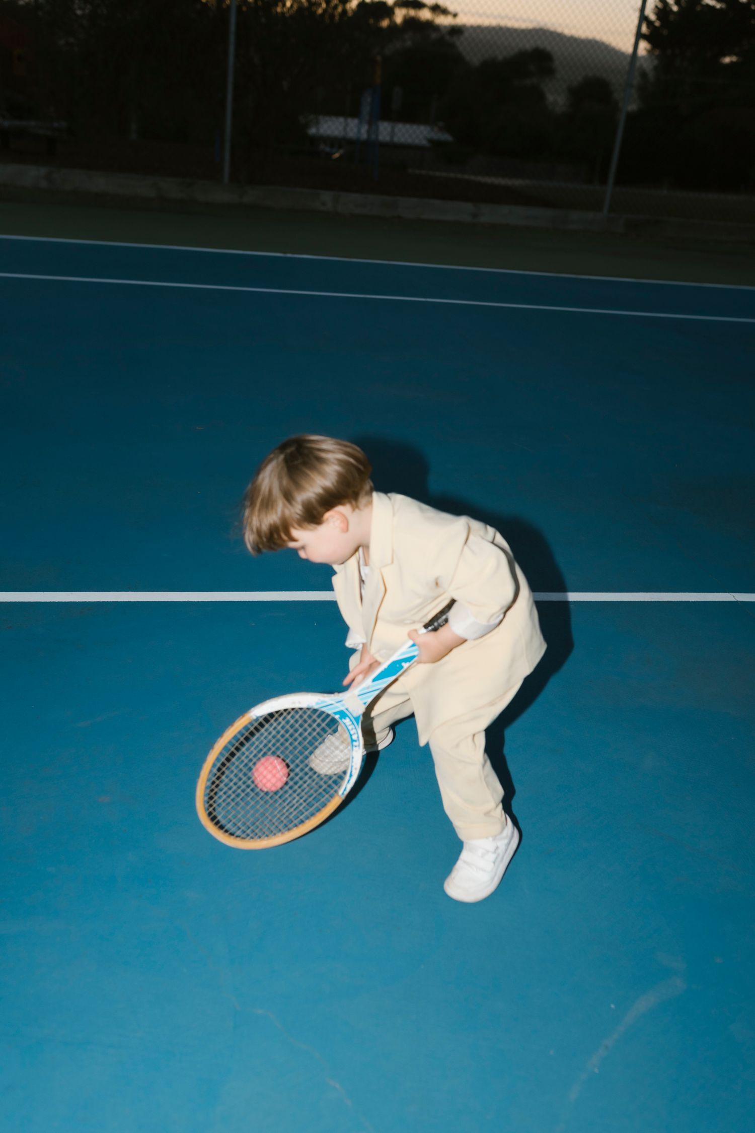 Young child in beige outfit playing tennis on a blue court, holding a racket and hitting a pink ball.