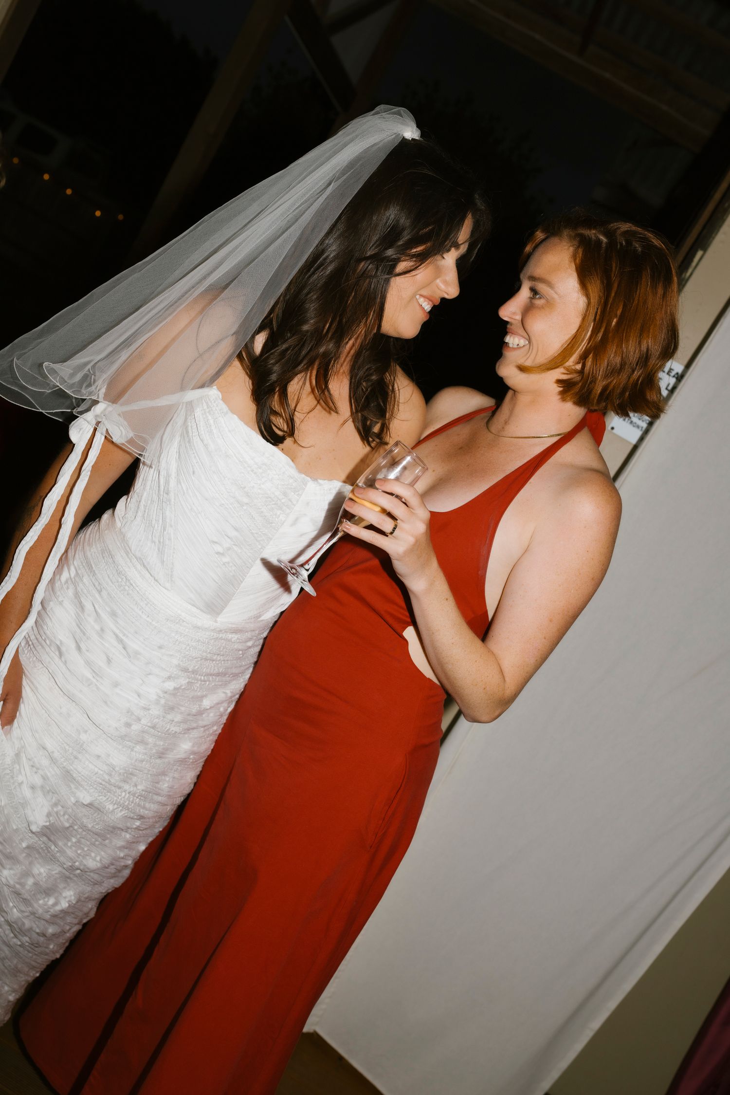 Bride in white gown and veil shares a champagne toast with bridesmaid in red dress at wedding reception.