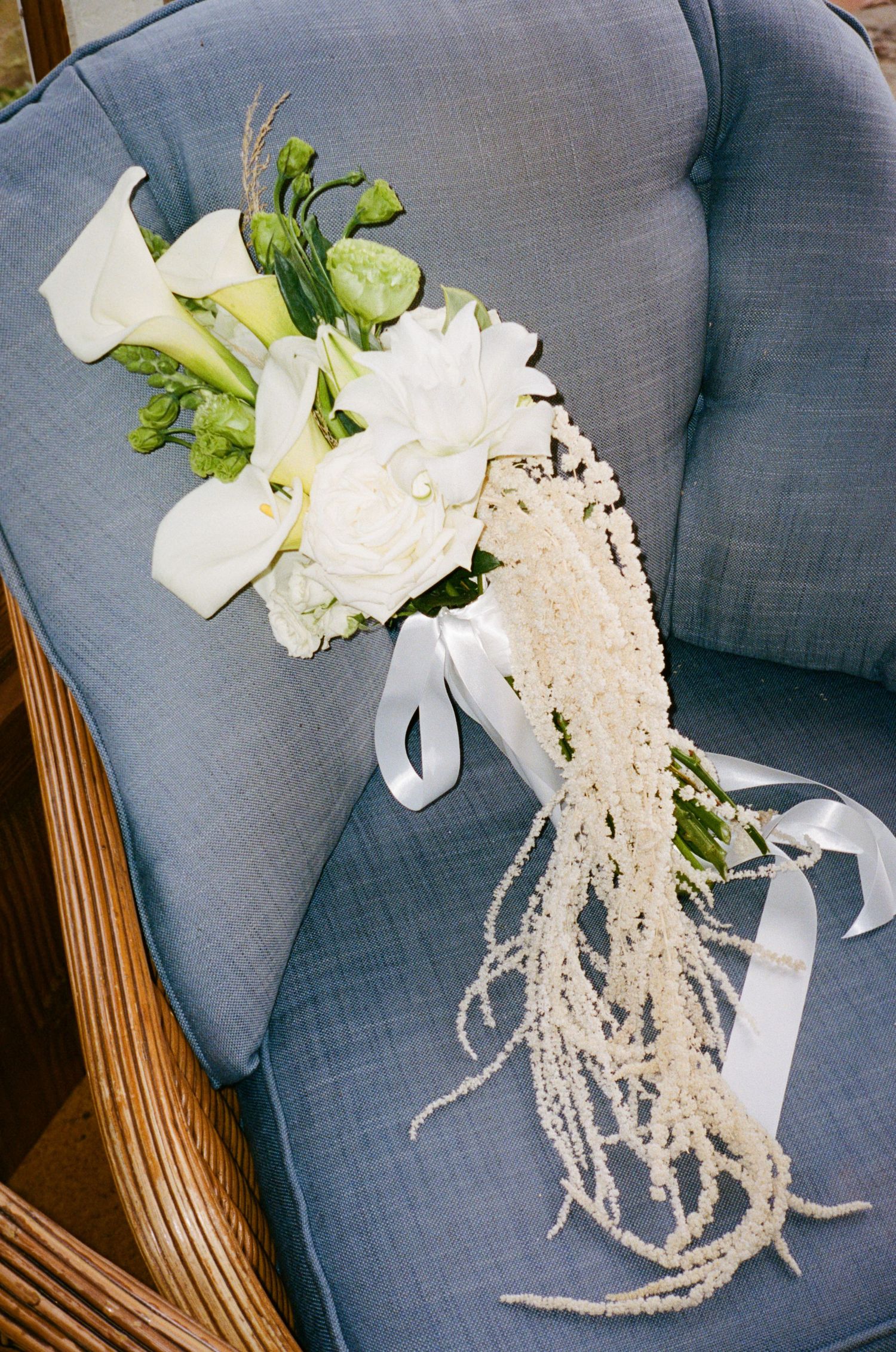 Elegant white bridal bouquet with calla lilies, roses, and cascading amaranthus on a blue linen chair.