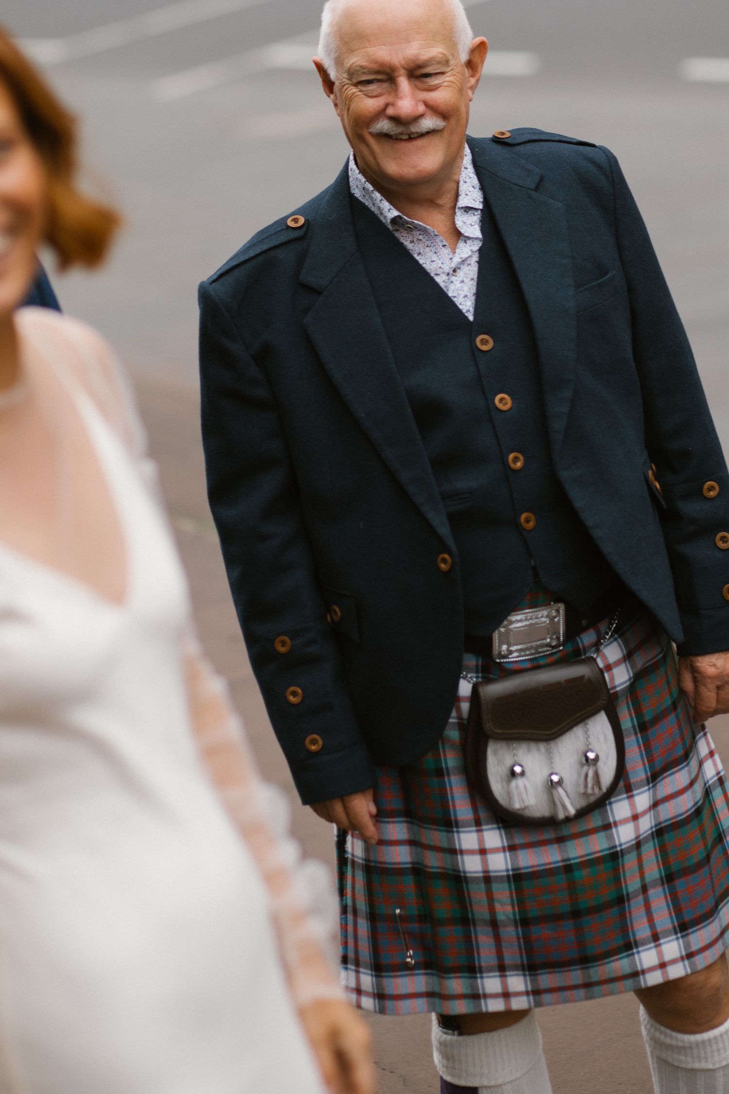 Smiling older man in traditional Scottish kilt and navy blazer with sporran at a wedding.