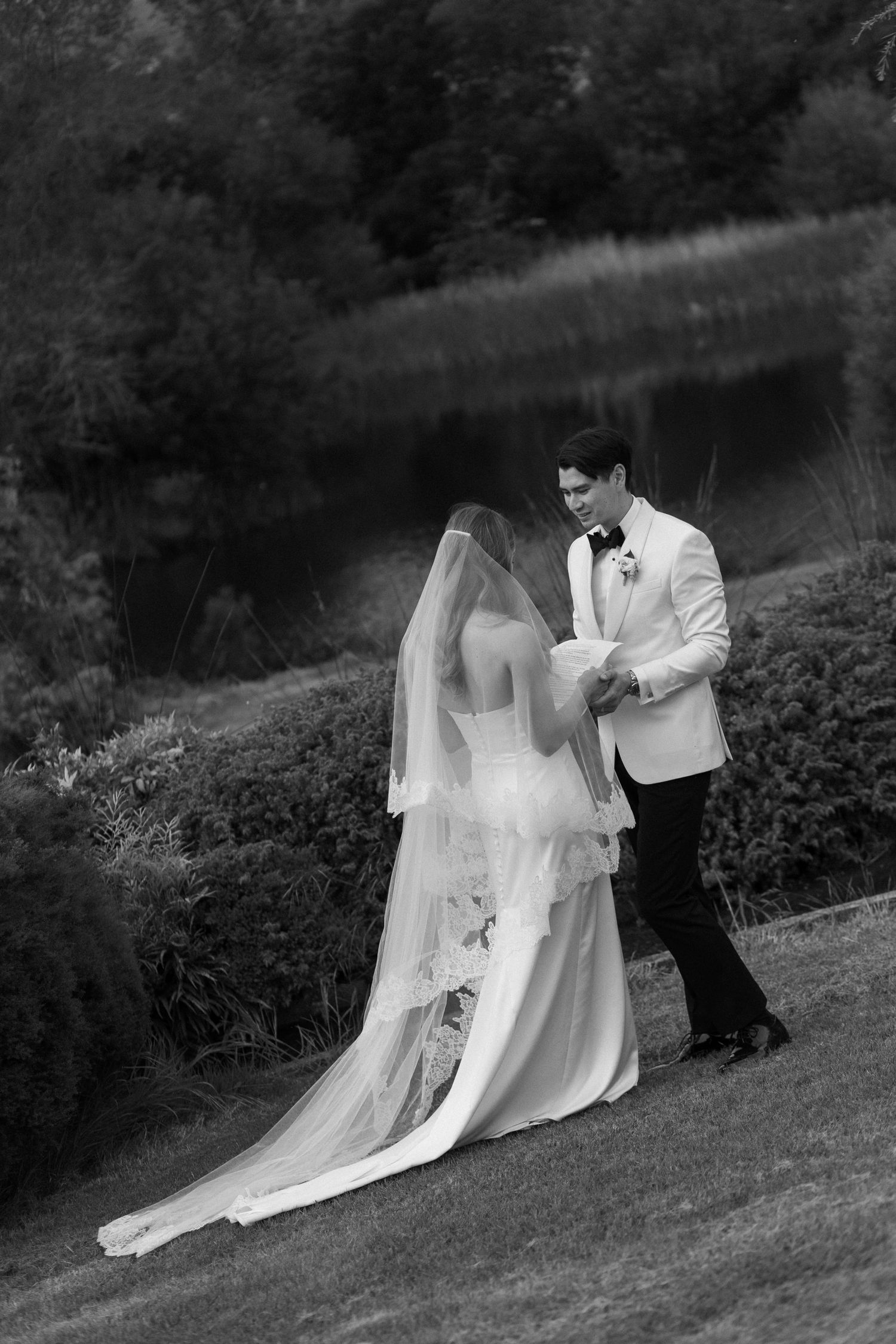 Black and white photo of groom reading vows to veiled bride in lush garden setting.
