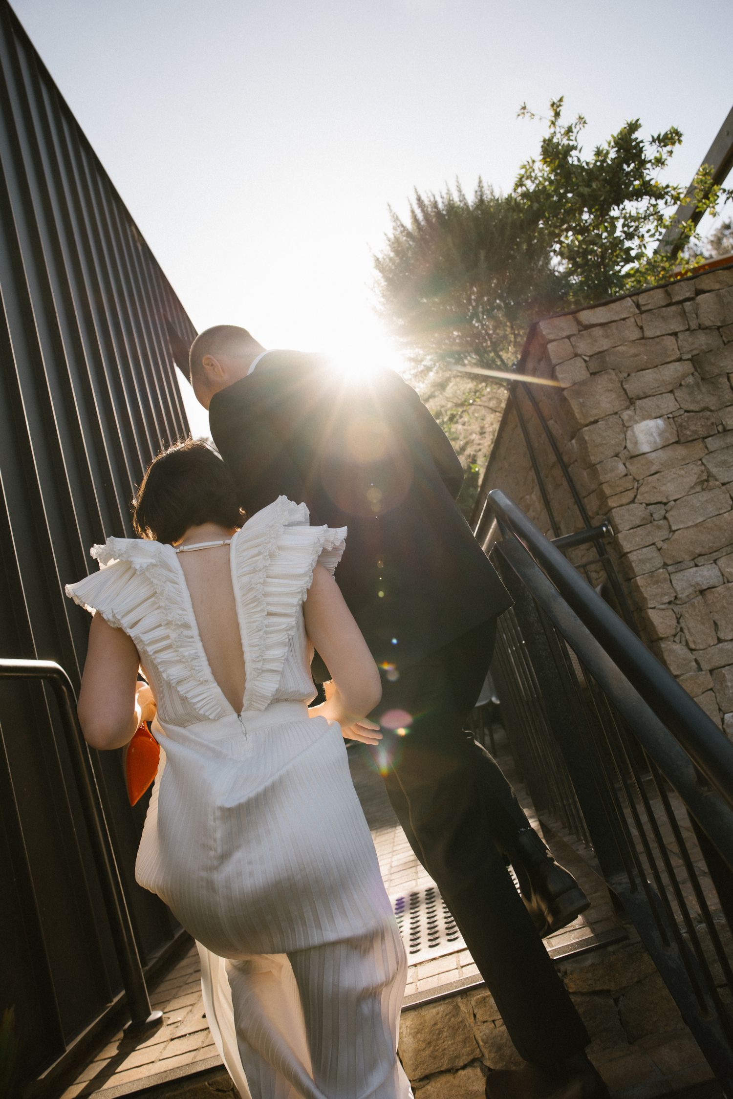 Bride and groom walking up sunlit outdoor stairs, bride in white ruffled backless gown.