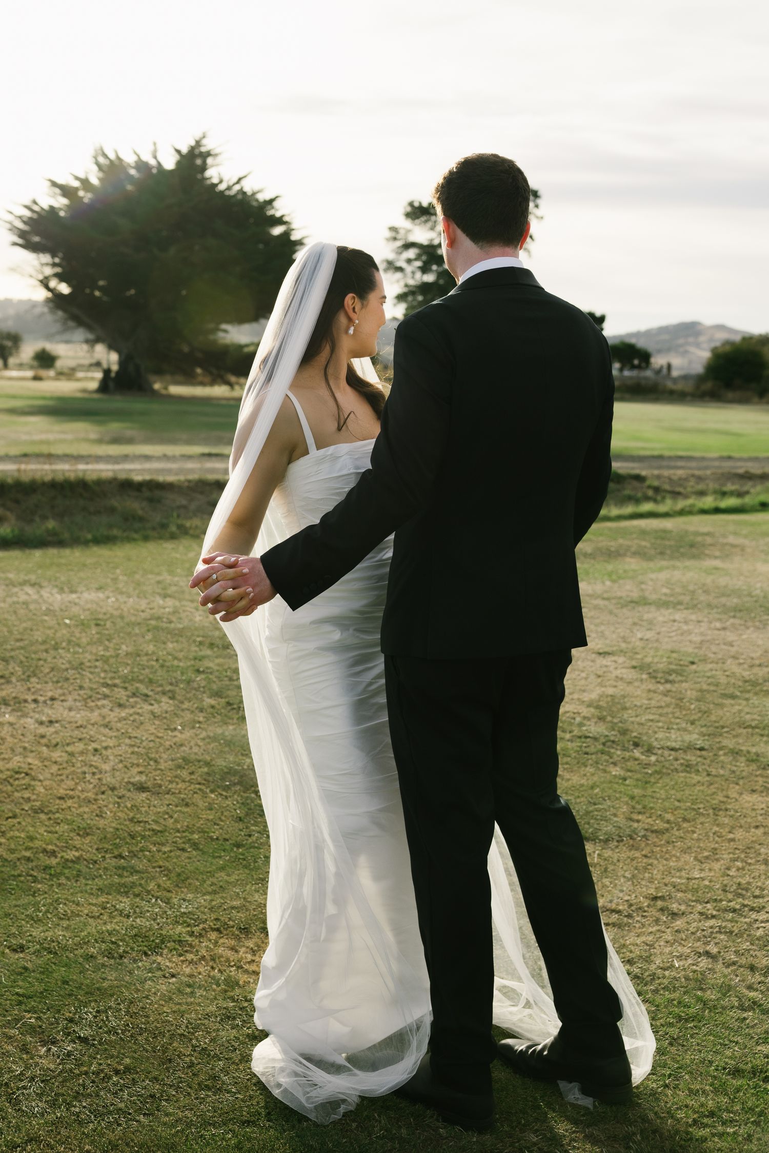 Bride in white gown and veil holding hands with groom in black suit on green lawn.