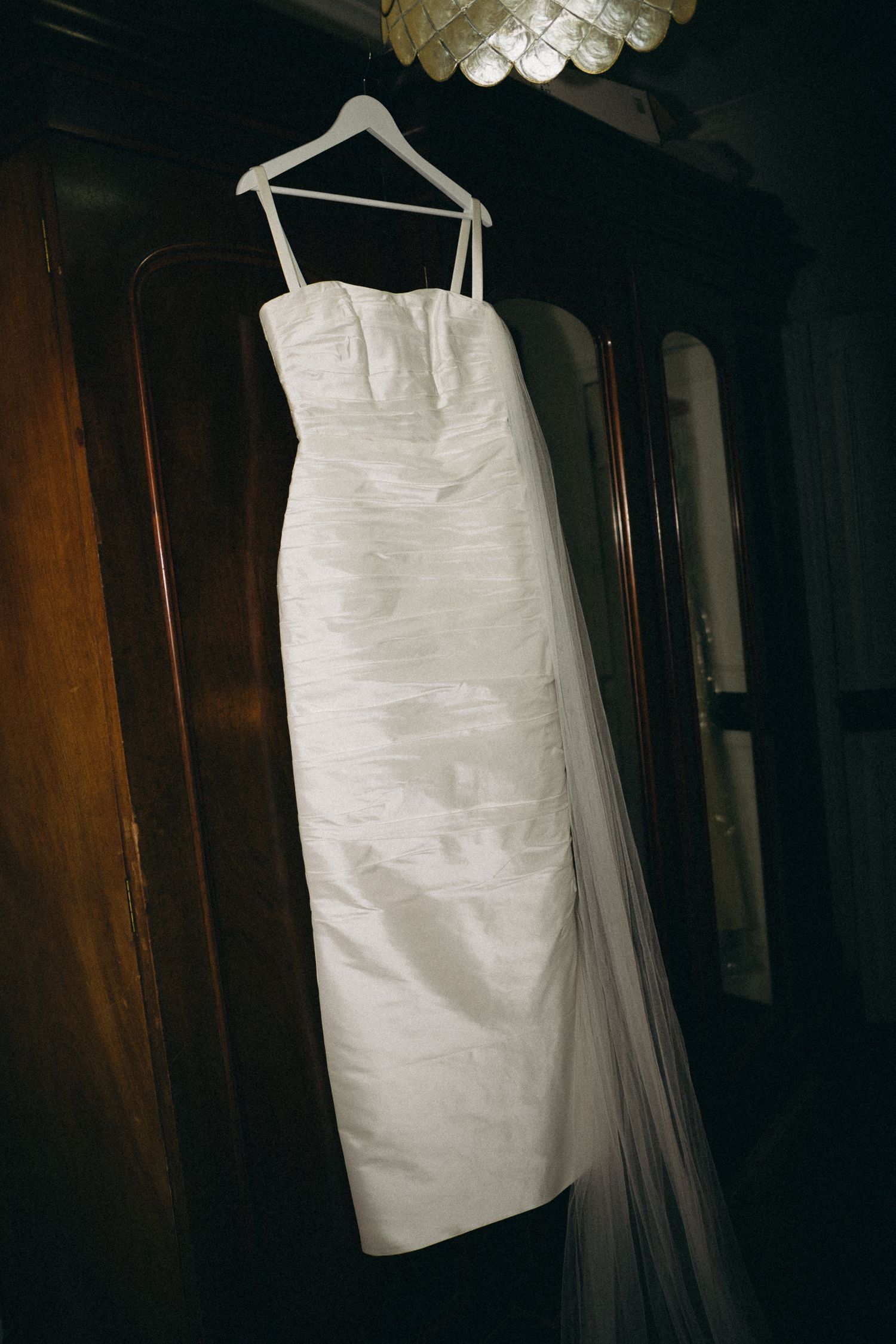 White strapless wedding dress with spaghetti straps and veil hanging on hanger against wooden wardrobe.