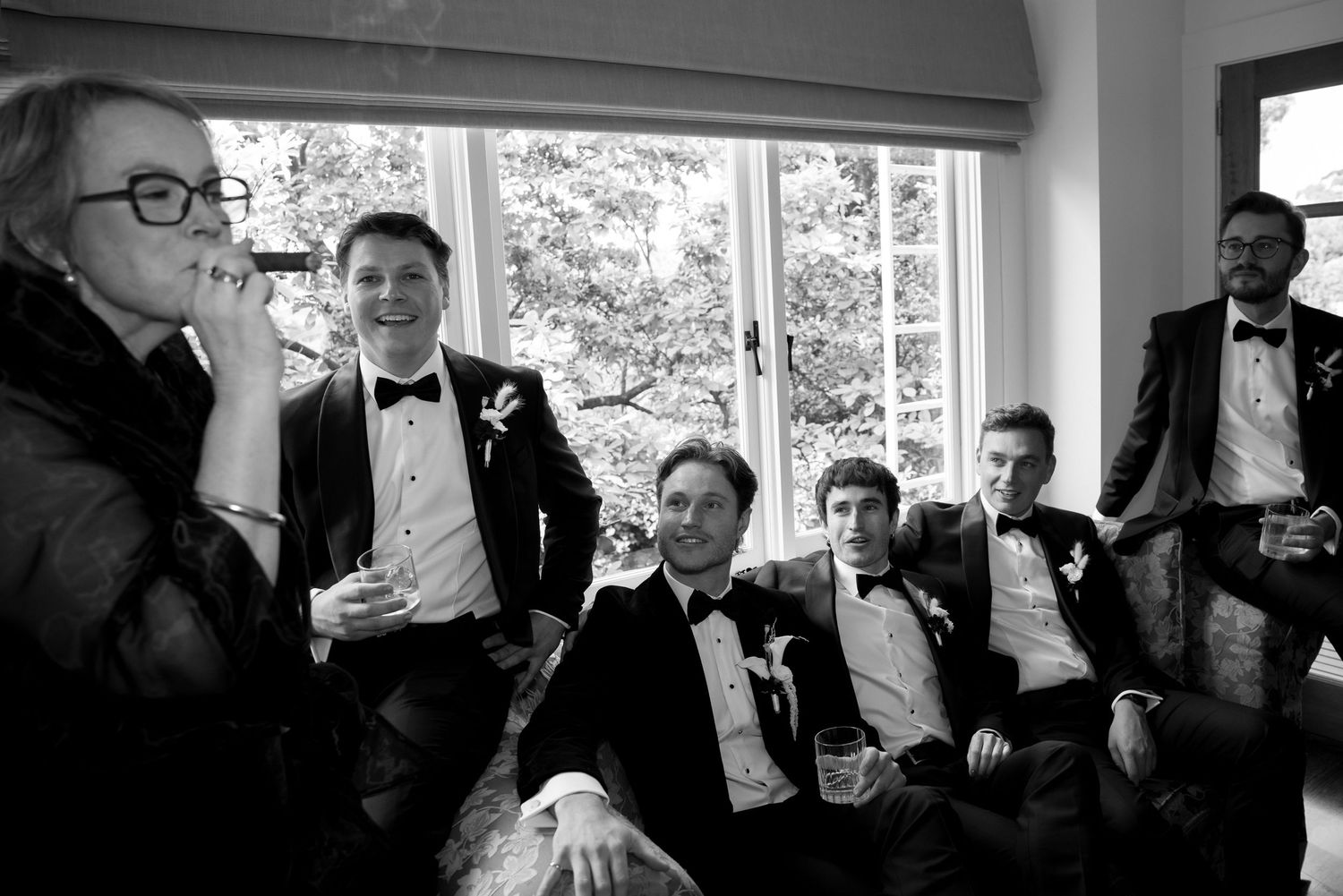 Groomsmen in black tuxedos relaxing and laughing together indoors before a wedding ceremony in black and white photo.