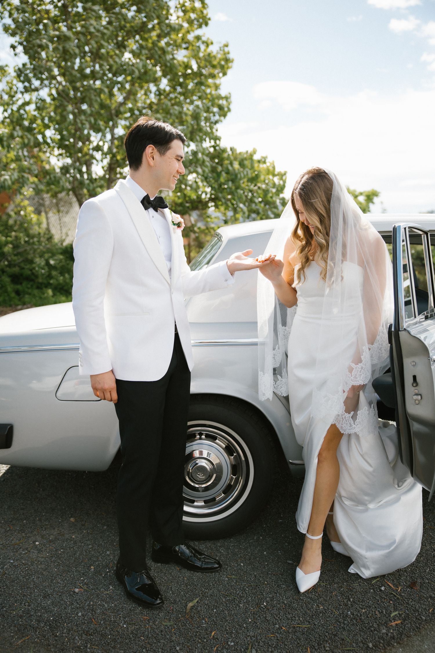 Groom in white tuxedo jacket helping bride in lace veil exit vintage white classic car.