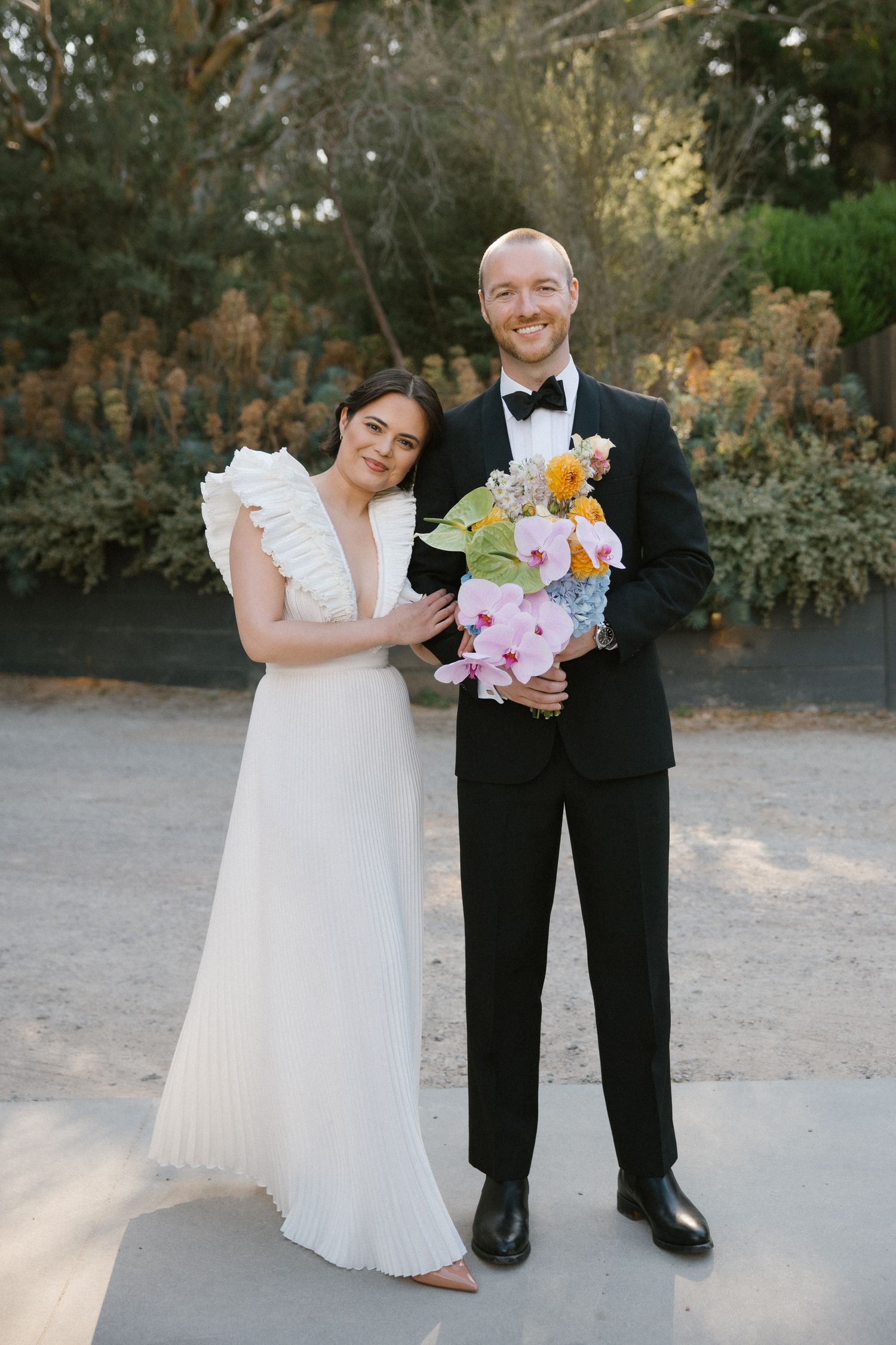 Smiling bride and groom outdoors holding colorful bridal bouquet with orchids and tropical flowers.
