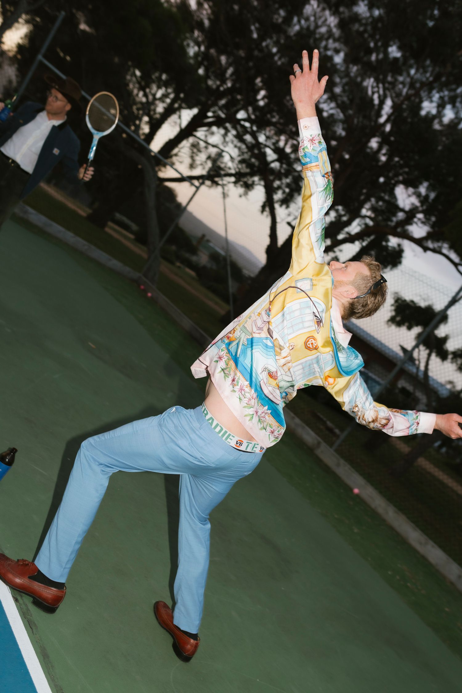 A man in a colorful printed shirt and blue pants stretches dramatically on an outdoor tennis court.
