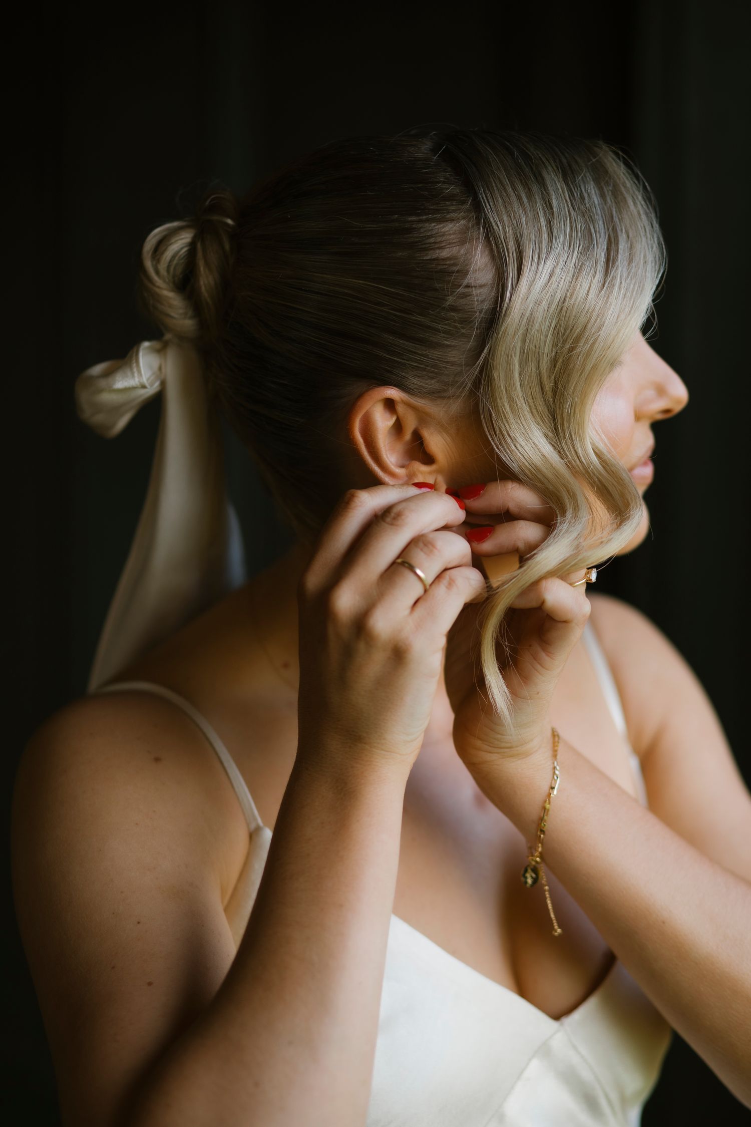 Side profile of a blonde bride with a white bow updo putting on earrings in a satin bridal gown.
