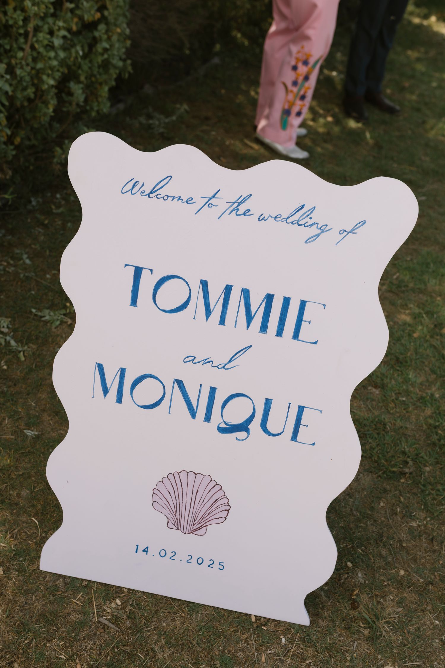 White wavy wedding welcome sign for Tommie and Monique, dated 14.02.2025, with blue text and shell illustration.