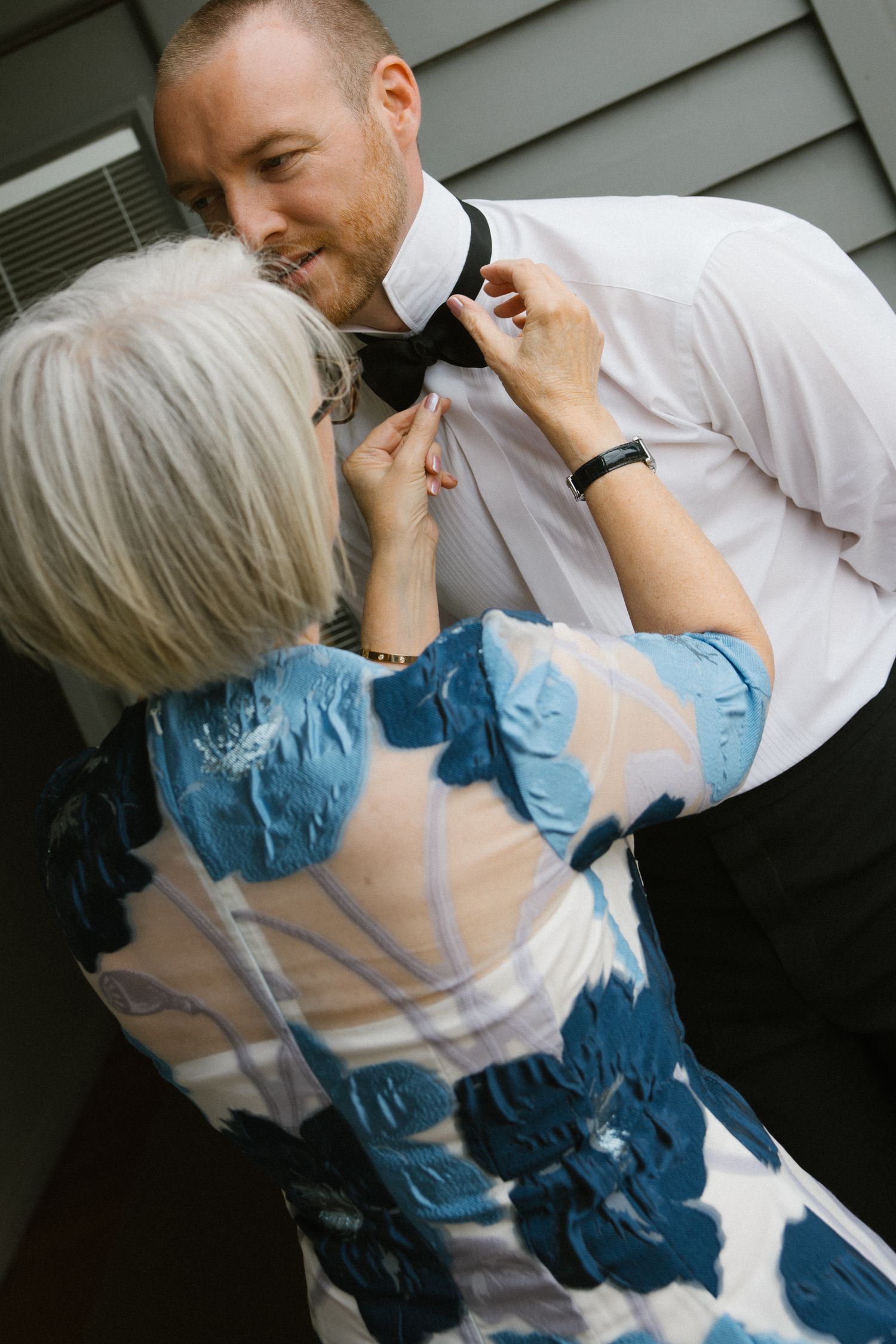 Older woman in blue floral dress adjusting groom's black bow tie before wedding ceremony.