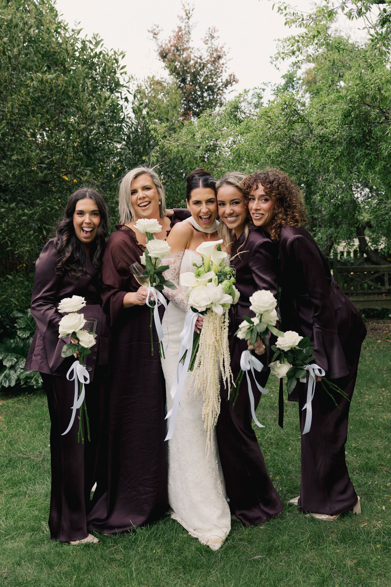 Bride in white lace gown poses with three bridesmaids in dark purple satin dresses holding white roses.